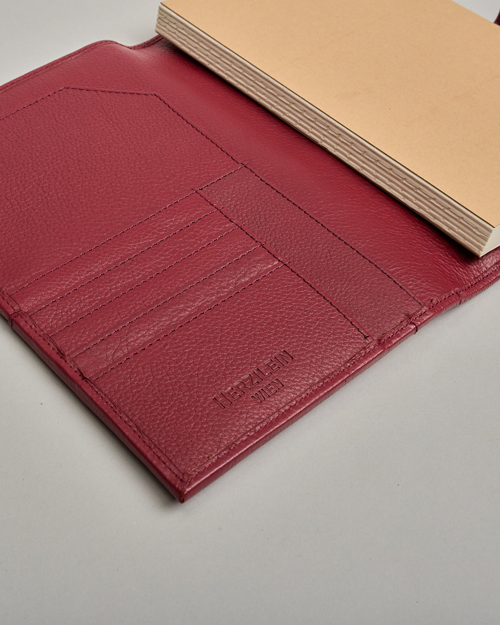 Bordeauxroter Leder Notizbucheinband mit leichter Narbund. Der Einband hat außen ein flaches Fach in das ein Stift und das Handy gesteckt werden können. Das Notizbuch lässt sich seitlich mit einer Lasche und einem darunterliegenden Druckknopf verschließen. Innen bietet das Notizbuch rechts Platz um ein Buch einzustecken und links gibt es viele Fächer für weitere Notizzettel, Visitkarten und mehr. #6