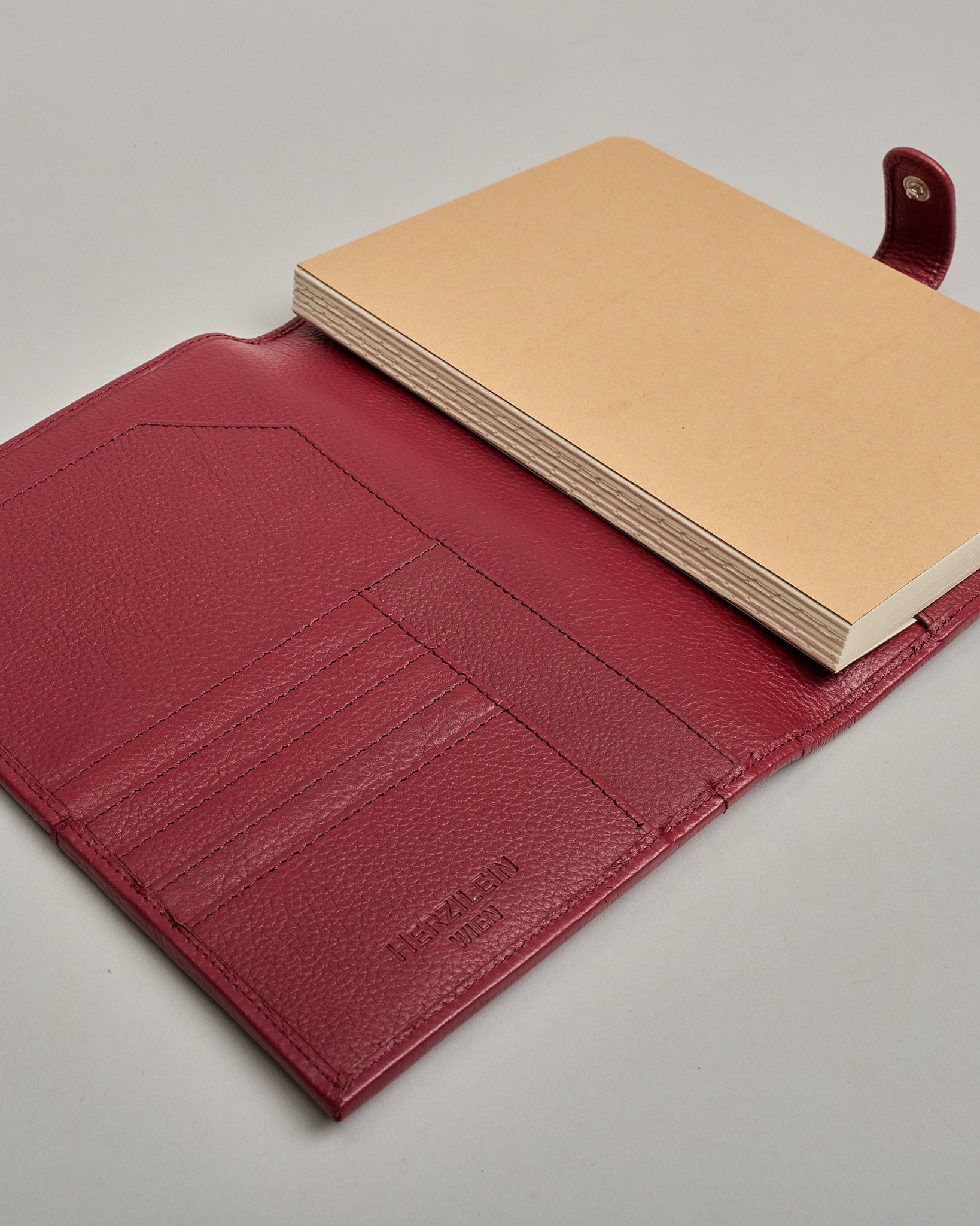 Bordeauxroter Leder Notizbucheinband mit leichter Narbund. Der Einband hat außen ein flaches Fach in das ein Stift und das Handy gesteckt werden können. Das Notizbuch lässt sich seitlich mit einer Lasche und einem darunterliegenden Druckknopf verschließen. Innen bietet das Notizbuch rechts Platz um ein Buch einzustecken und links gibt es viele Fächer für weitere Notizzettel, Visitkarten und mehr. #7