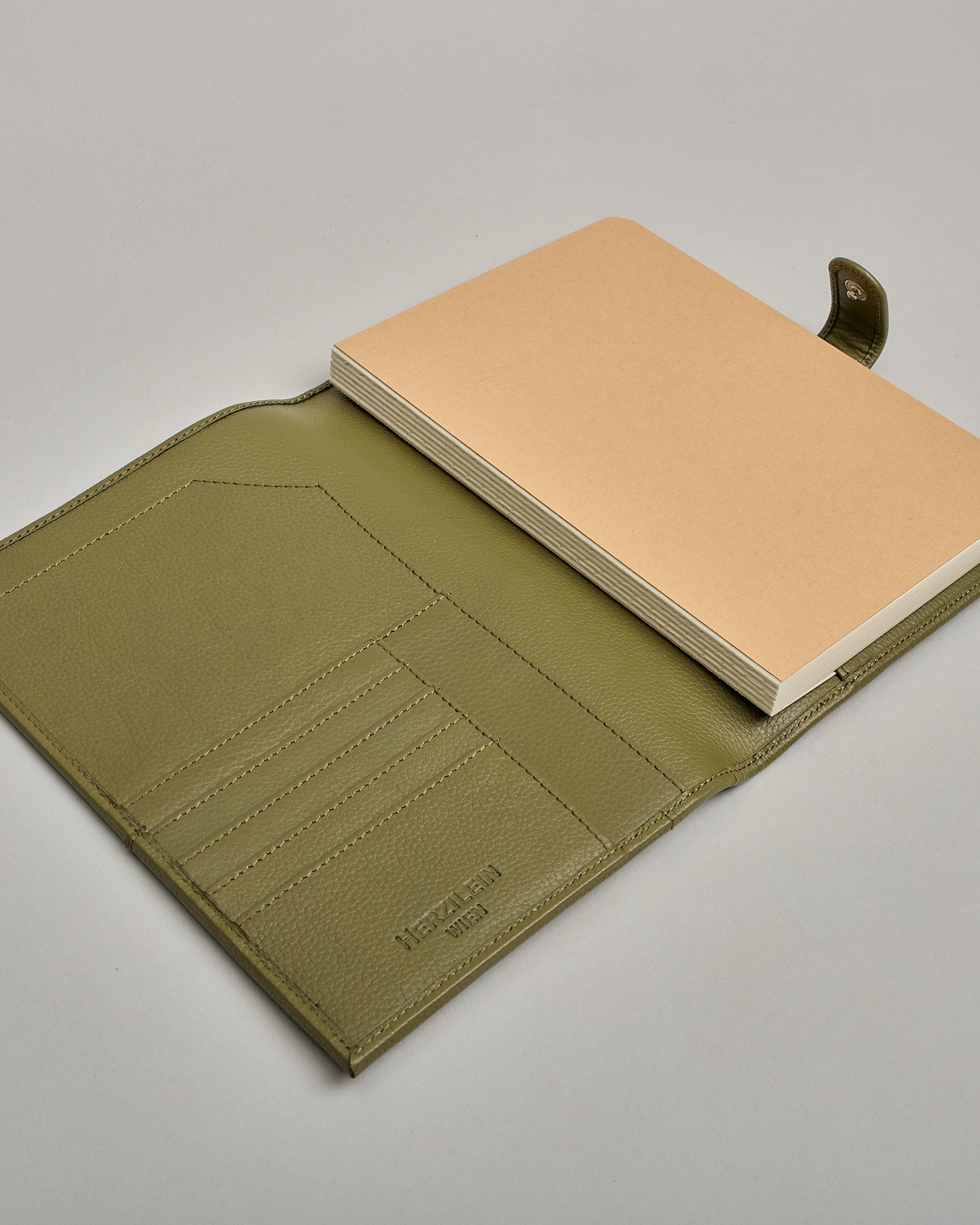 Grüner Leder Notizbucheinband mit leichter Narbund. Der Einband hat außen ein flaches Fach in das ein Stift und das Handy gesteckt werden können. Das Notizbuch lässt sich seitlich mit einer Lasche und einem darunterliegenden Druckknopf verschließen. Innen bietet das Notizbuch rechts Platz um ein Buch einzustecken und links gibt es viele Fächer für weitere Notizzettel, Visitkarten und mehr. #7