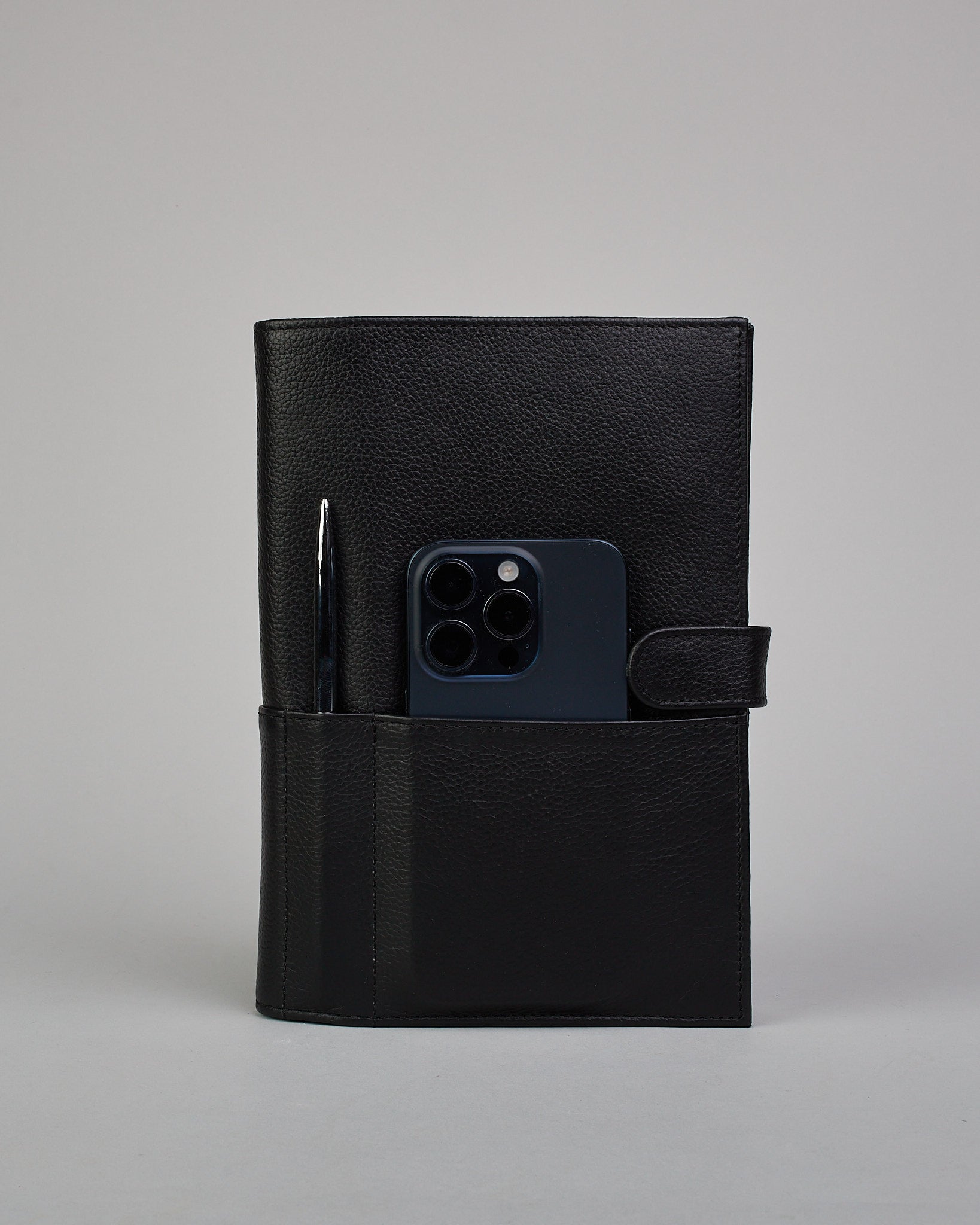 Schwarzer Leder Notizbucheinband mit leichter Narbund. Der Einband hat außen ein flaches Fach in das ein Stift und das Handy gesteckt werden können. Das Notizbuch lässt sich seitlich mit einer Lasche und einem darunterliegenden Druckknopf verschließen. Innen bietet das Notizbuch rechts Platz um ein Buch einzustecken und links gibt es viele Fächer für weitere Notizzettel, Visitkarten und mehr. #4