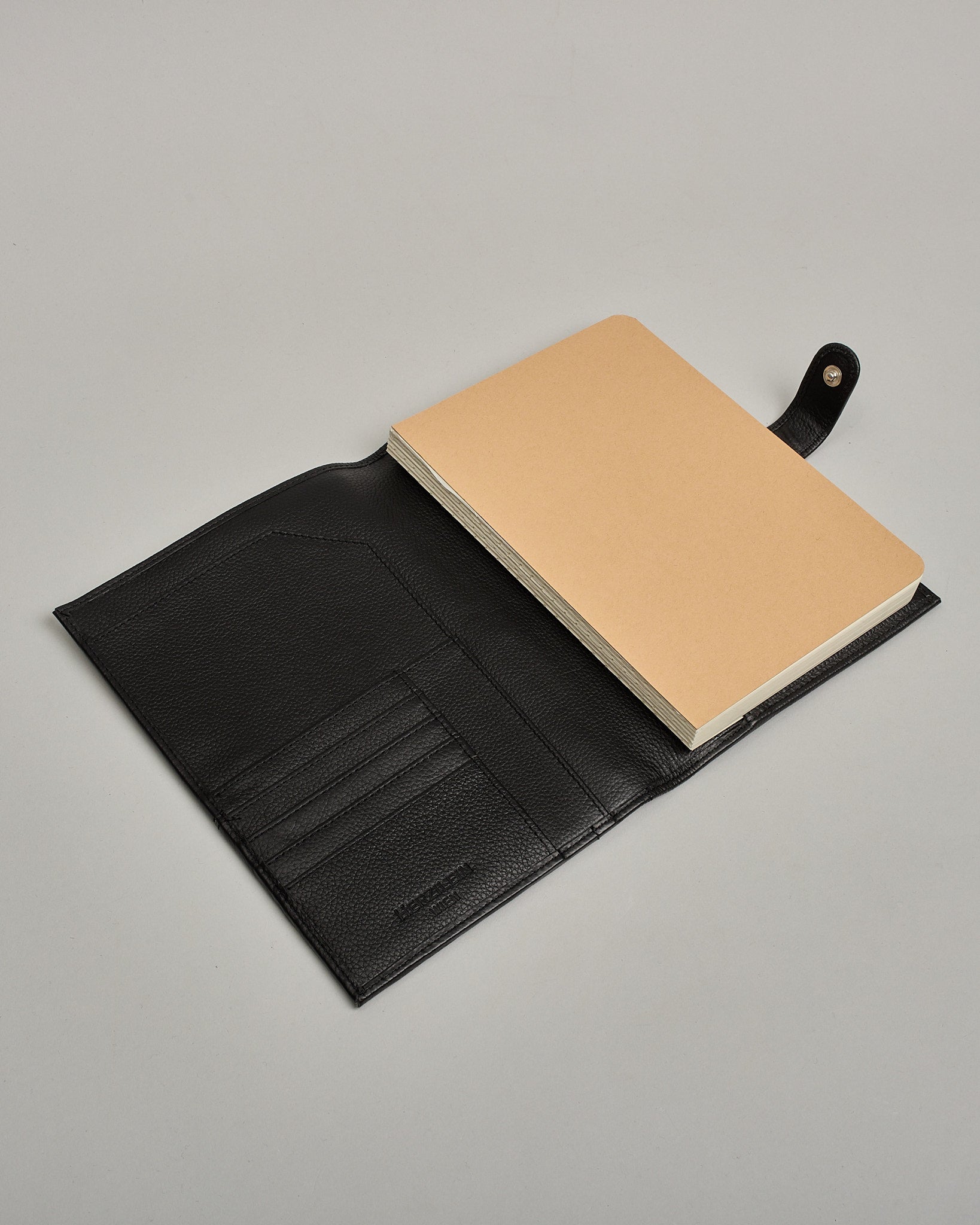 Schwarzer Leder Notizbucheinband mit leichter Narbund. Der Einband hat außen ein flaches Fach in das ein Stift und das Handy gesteckt werden können. Das Notizbuch lässt sich seitlich mit einer Lasche und einem darunterliegenden Druckknopf verschließen. Innen bietet das Notizbuch rechts Platz um ein Buch einzustecken und links gibt es viele Fächer für weitere Notizzettel, Visitkarten und mehr. #5