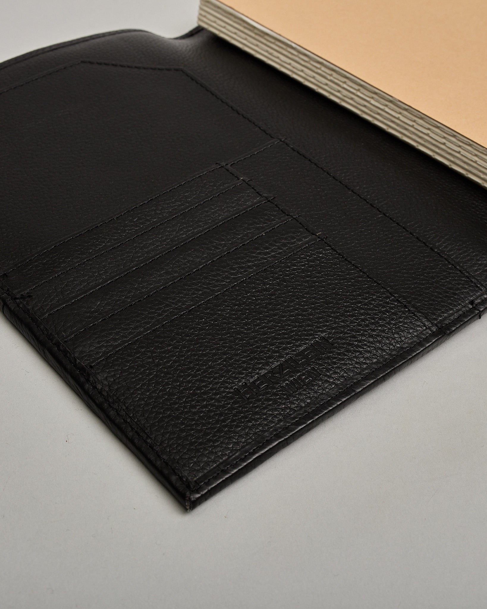 Schwarzer Leder Notizbucheinband mit leichter Narbund. Der Einband hat außen ein flaches Fach in das ein Stift und das Handy gesteckt werden können. Das Notizbuch lässt sich seitlich mit einer Lasche und einem darunterliegenden Druckknopf verschließen. Innen bietet das Notizbuch rechts Platz um ein Buch einzustecken und links gibt es viele Fächer für weitere Notizzettel, Visitkarten und mehr. #6