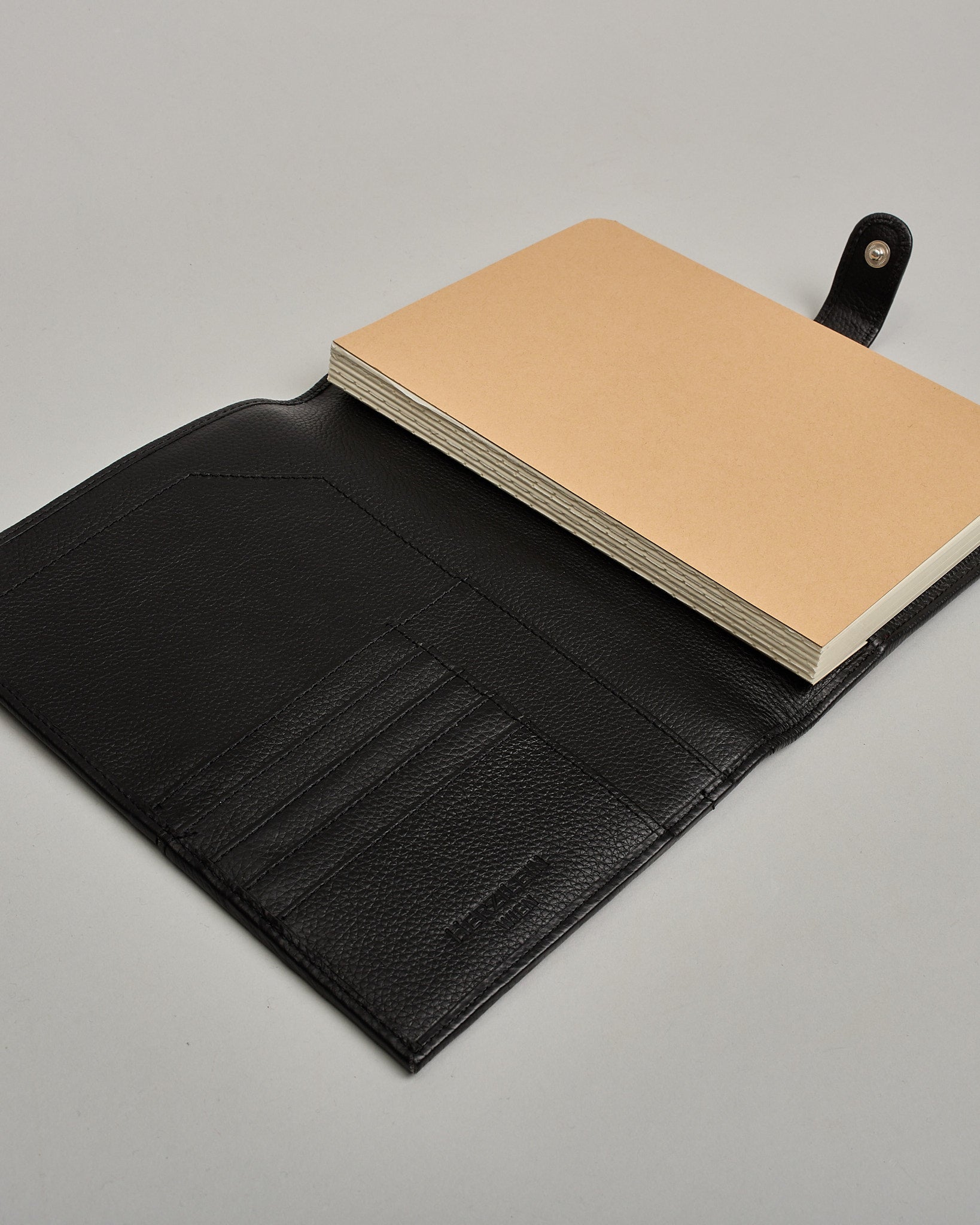 Schwarzer Leder Notizbucheinband mit leichter Narbund. Der Einband hat außen ein flaches Fach in das ein Stift und das Handy gesteckt werden können. Das Notizbuch lässt sich seitlich mit einer Lasche und einem darunterliegenden Druckknopf verschließen. Innen bietet das Notizbuch rechts Platz um ein Buch einzustecken und links gibt es viele Fächer für weitere Notizzettel, Visitkarten und mehr. #7