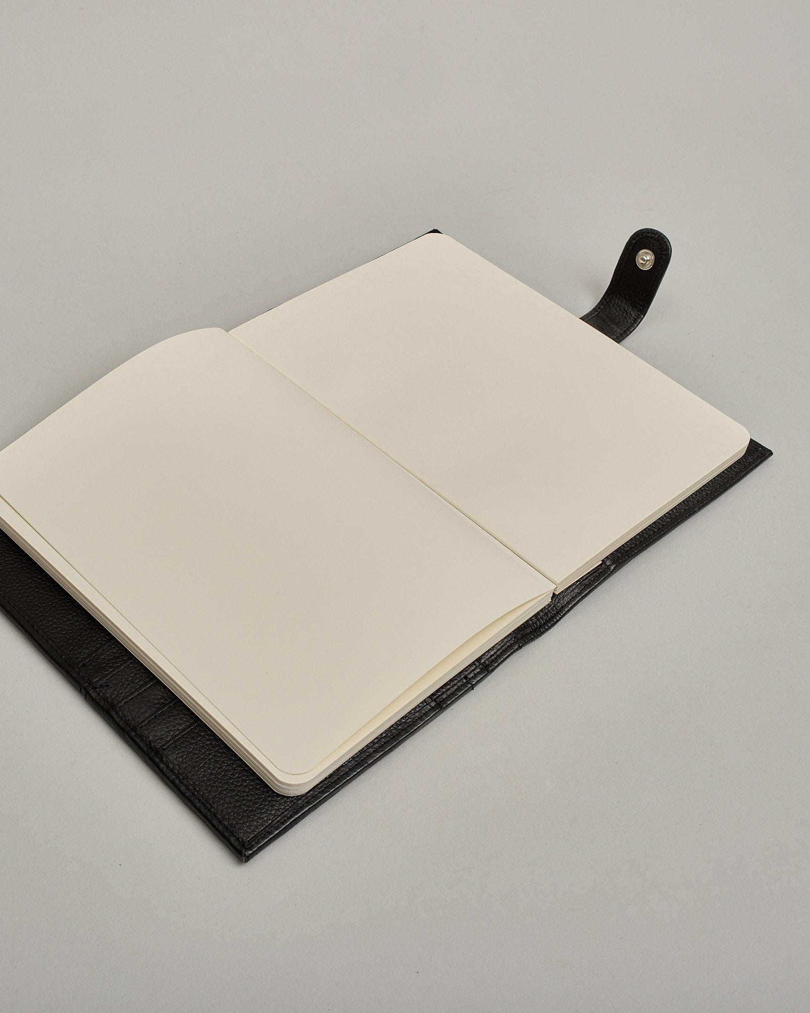 Schwarzer Leder Notizbucheinband mit leichter Narbund. Der Einband hat außen ein flaches Fach in das ein Stift und das Handy gesteckt werden können. Das Notizbuch lässt sich seitlich mit einer Lasche und einem darunterliegenden Druckknopf verschließen. Innen bietet das Notizbuch rechts Platz um ein Buch einzustecken und links gibt es viele Fächer für weitere Notizzettel, Visitkarten und mehr. #8