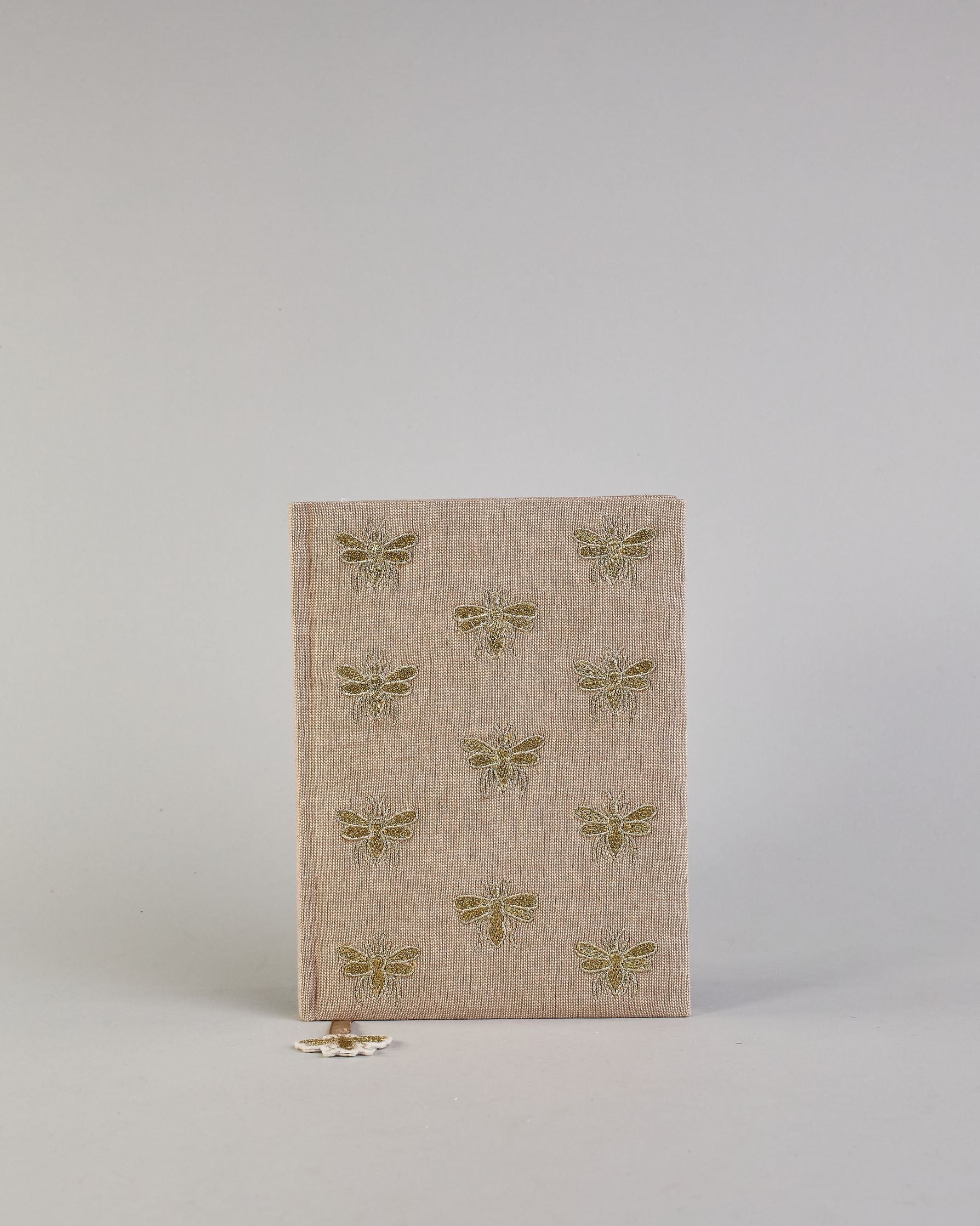 Beiges Notizbuch mti Baumwolleinband und vielen gestickten goldenen Bienen in regelmäßigen Abständen auf dem Einband. Lesezeichen mit goldener Biene als Anhänger. Linierte Seiten innen. #1