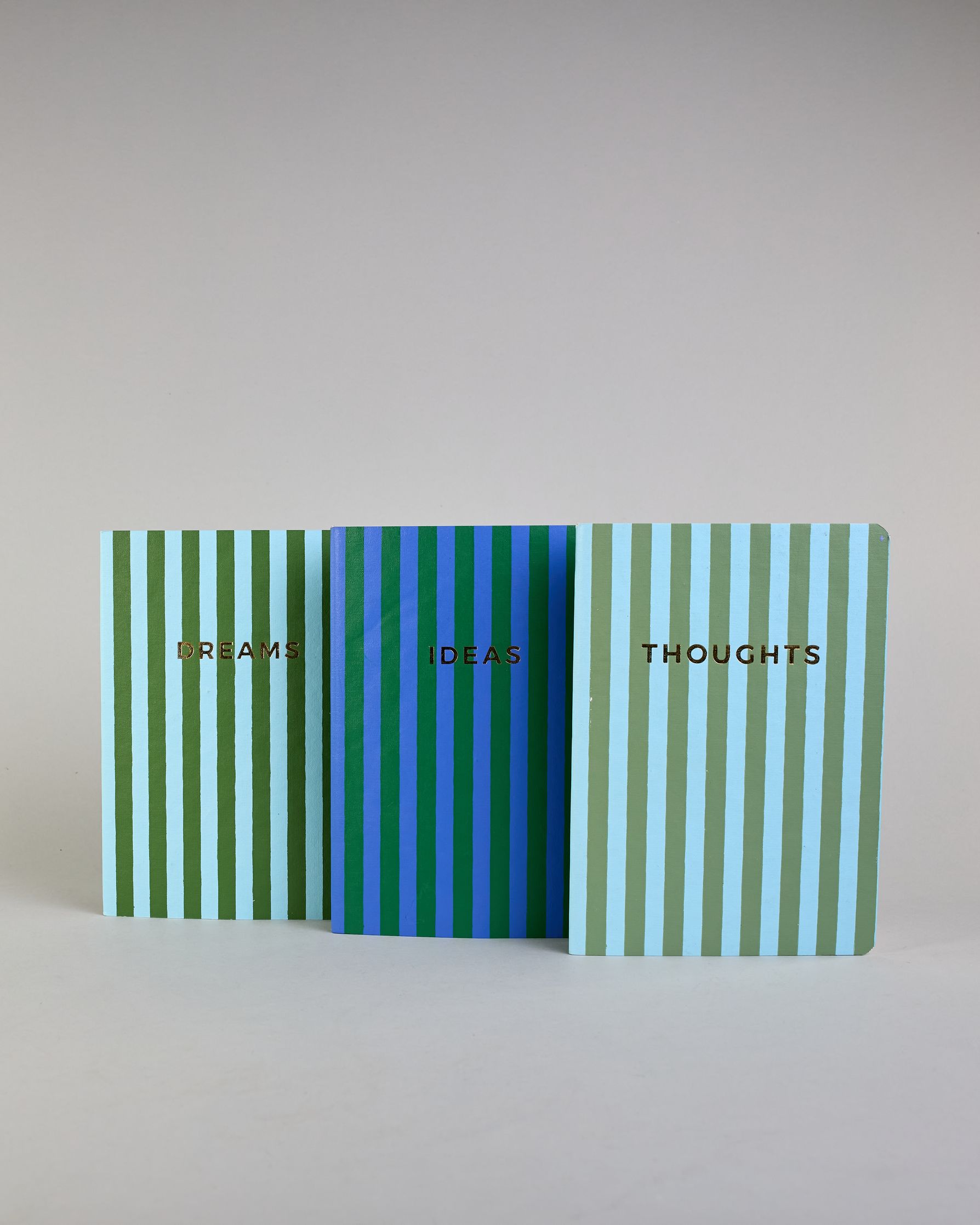 Set aus drei A5-Heften mit recyceltem Baumwolleinband. Die Hefte sind mit senkrechten Streifen in Blau- und Grüntönen gestaltet und zeigen auf dem Cover jeweils ein goldener Spruch: Dreams, Thoughts und Ideas. 30 linierte Seiten innen. #1