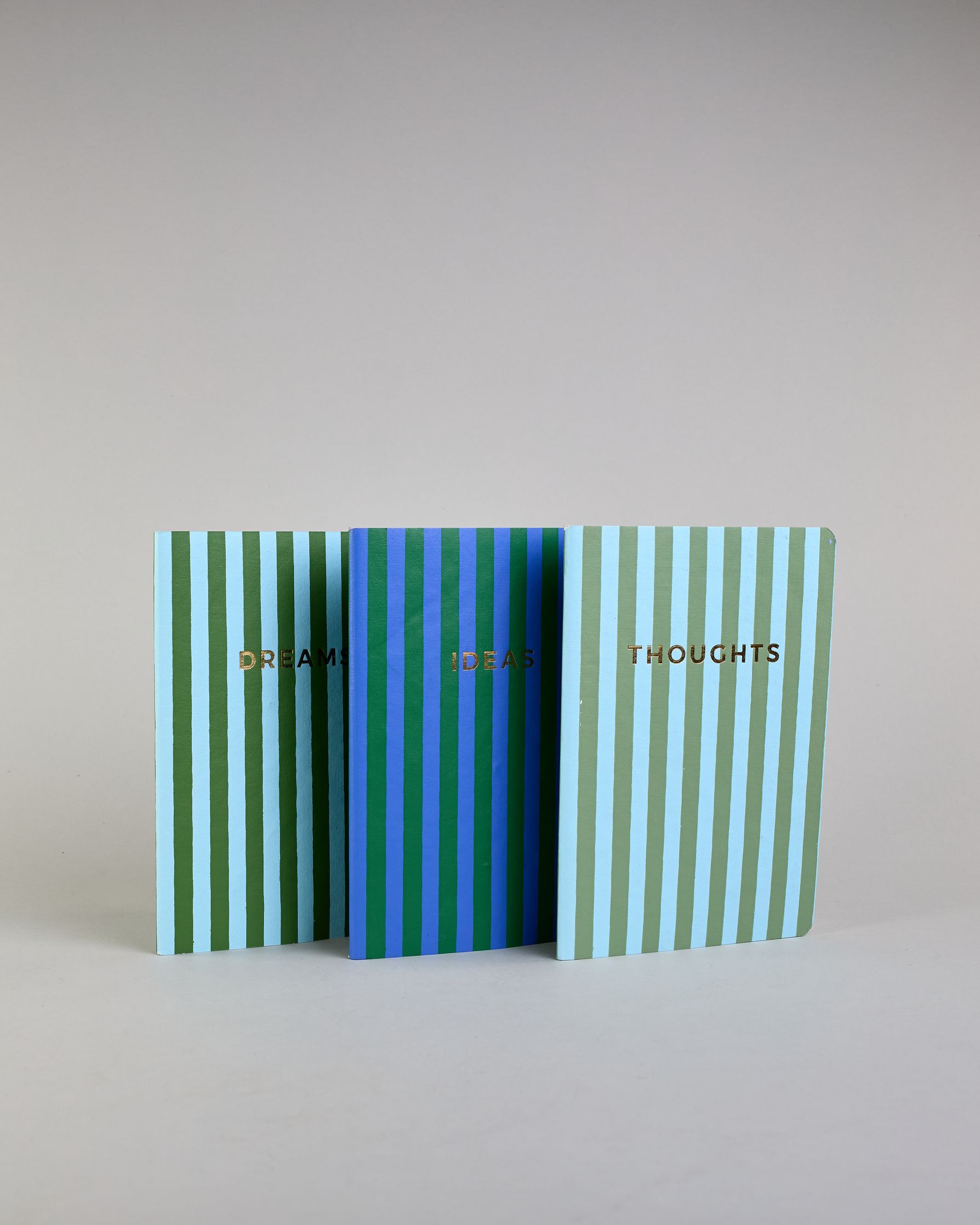 Set aus drei A5-Heften mit recyceltem Baumwolleinband. Die Hefte sind mit senkrechten Streifen in Blau- und Grüntönen gestaltet und zeigen auf dem Cover jeweils ein goldener Spruch: Dreams, Thoughts und Ideas. 30 linierte Seiten innen. #2