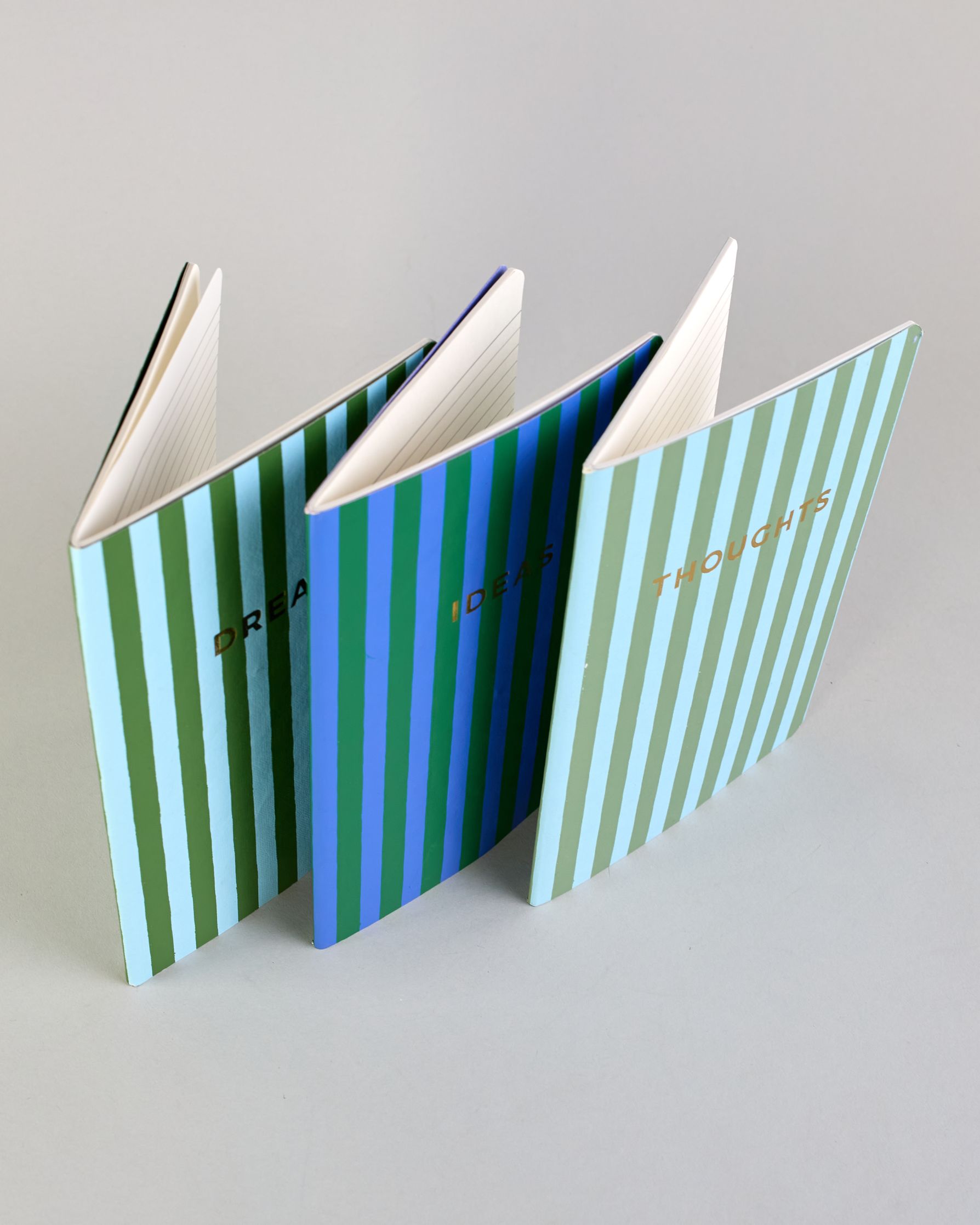 Set aus drei A5-Heften mit recyceltem Baumwolleinband. Die Hefte sind mit senkrechten Streifen in Blau- und Grüntönen gestaltet und zeigen auf dem Cover jeweils ein goldener Spruch: Dreams, Thoughts und Ideas. 30 linierte Seiten innen. #11