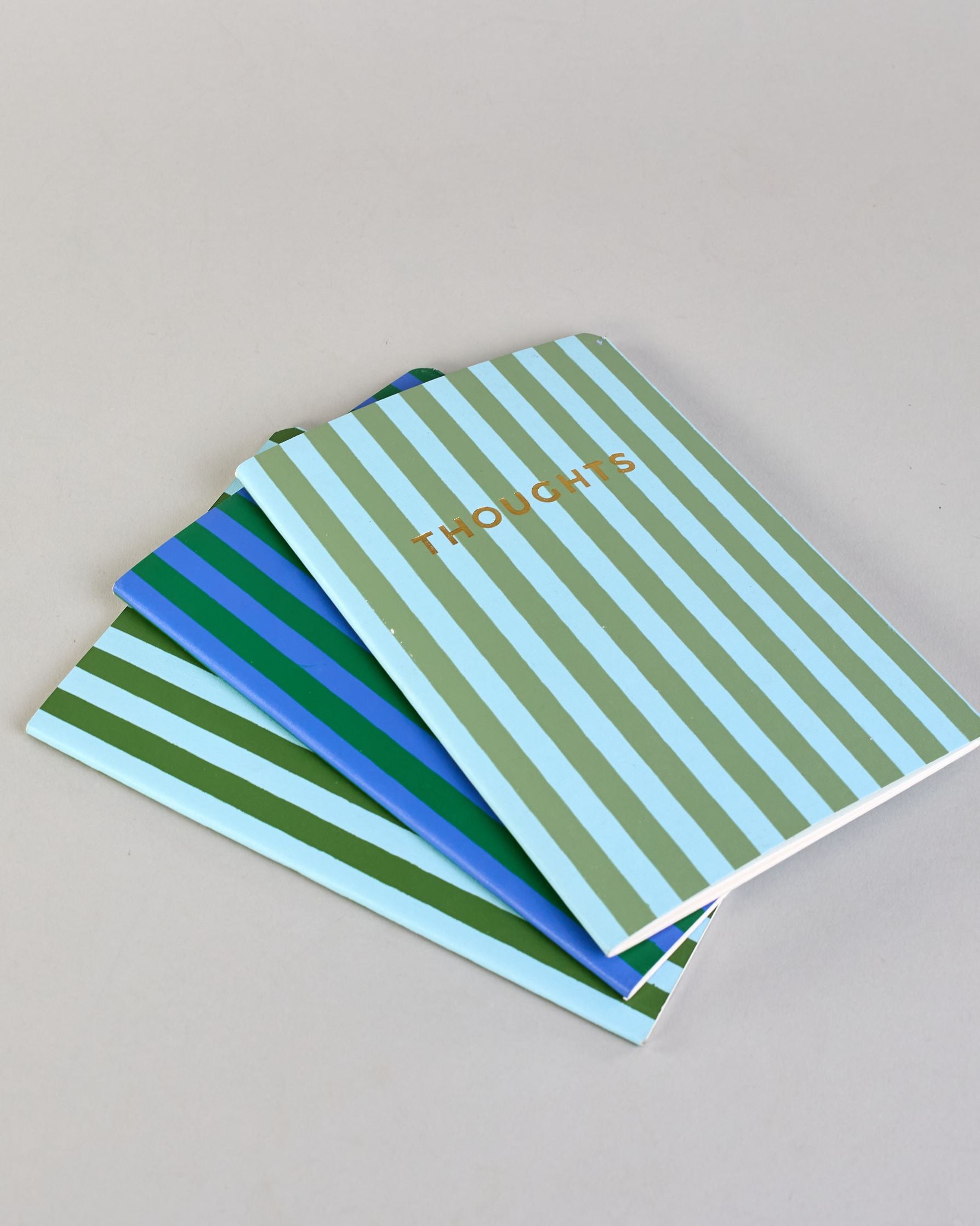 Set aus drei A5-Heften mit recyceltem Baumwolleinband. Die Hefte sind mit senkrechten Streifen in Blau- und Grüntönen gestaltet und zeigen auf dem Cover jeweils ein goldener Spruch: Dreams, Thoughts und Ideas. 30 linierte Seiten innen. #10