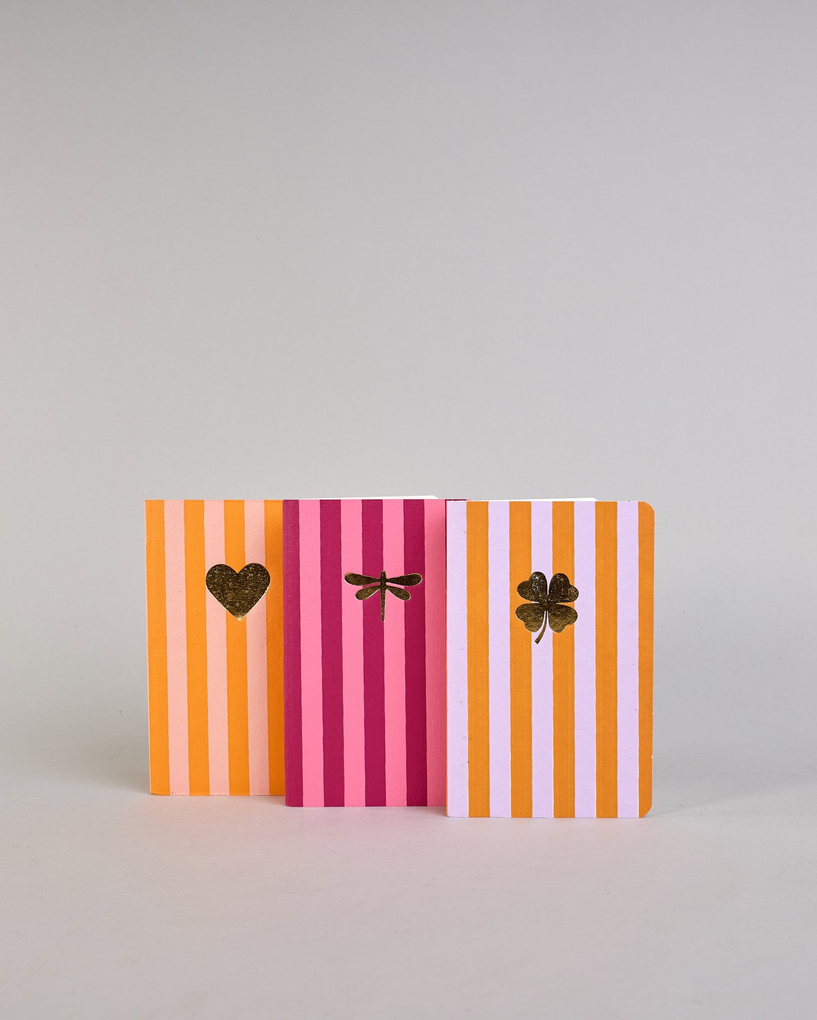 Set aus drei A6-Heften mit recyceltem Baumwolleinband. Die Hefte sind mit senkrechten Streifen in Blau- und Grüntönen gestaltet und zeigen auf dem Cover jeweils ein goldenes Motiv: ein Herz, ein Kleeblatt und eine Libelle. 30 glatte Seiten innen. #1