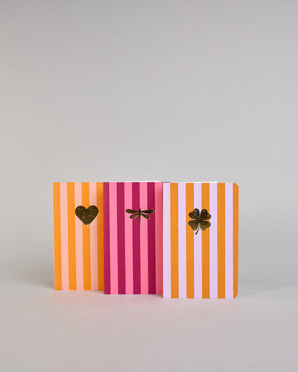 Set aus drei A6-Heften mit recyceltem Baumwolleinband. Die Hefte sind mit senkrechten Streifen in Blau- und Grüntönen gestaltet und zeigen auf dem Cover jeweils ein goldenes Motiv: ein Herz, ein Kleeblatt und eine Libelle. 30 glatte Seiten innen. #1