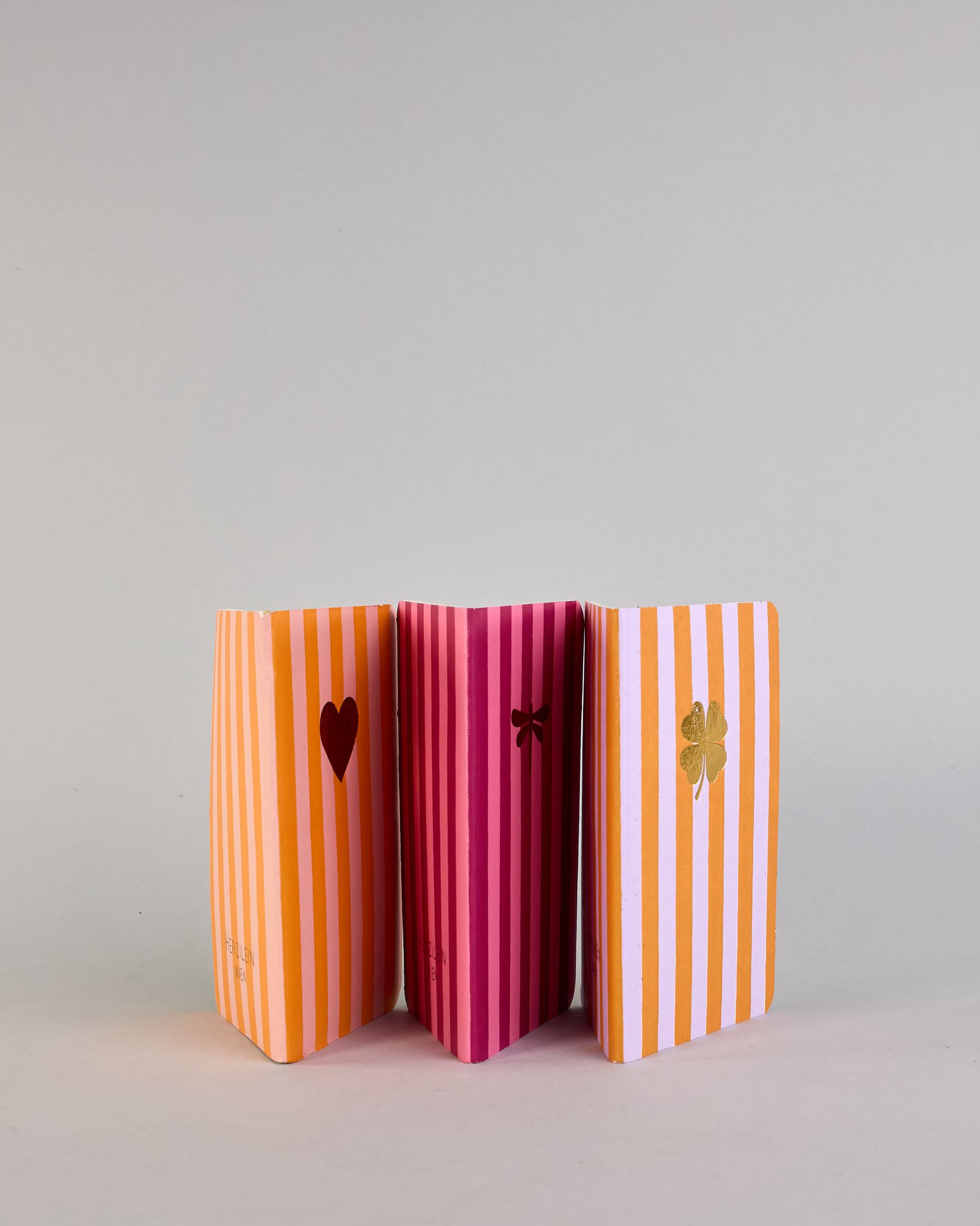 Set aus drei A6-Heften mit recyceltem Baumwolleinband. Die Hefte sind mit senkrechten Streifen in Blau- und Grüntönen gestaltet und zeigen auf dem Cover jeweils ein goldenes Motiv: ein Herz, ein Kleeblatt und eine Libelle. 30 glatte Seiten innen. #3