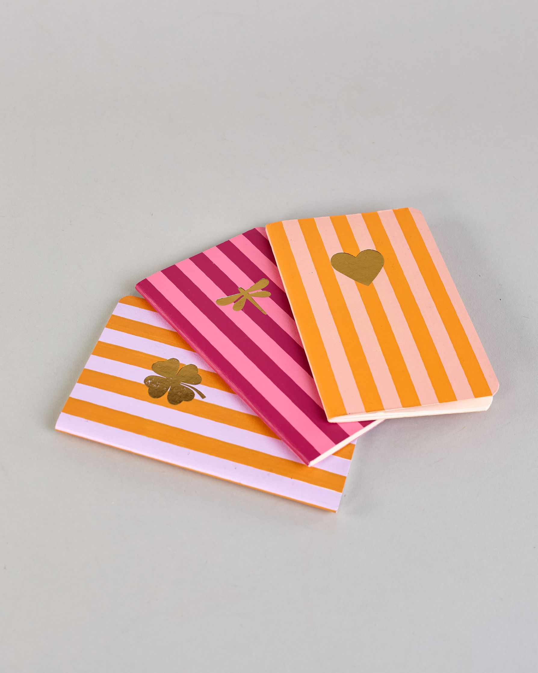 Set aus drei A6-Heften mit recyceltem Baumwolleinband. Die Hefte sind mit senkrechten Streifen in Blau- und Grüntönen gestaltet und zeigen auf dem Cover jeweils ein goldenes Motiv: ein Herz, ein Kleeblatt und eine Libelle. 30 glatte Seiten innen. #10