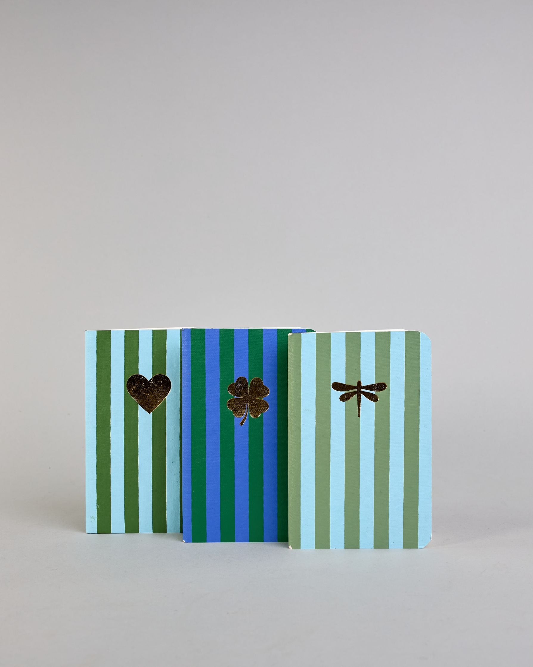 Set aus drei A6-Heften mit recyceltem Baumwolleinband. Die Hefte sind mit senkrechten Streifen in Blau- und Grüntönen gestaltet und zeigen auf dem Cover jeweils ein goldenes Motiv: ein Herz, ein Kleeblatt und eine Libelle. 30 glatte Seiten innen. #1