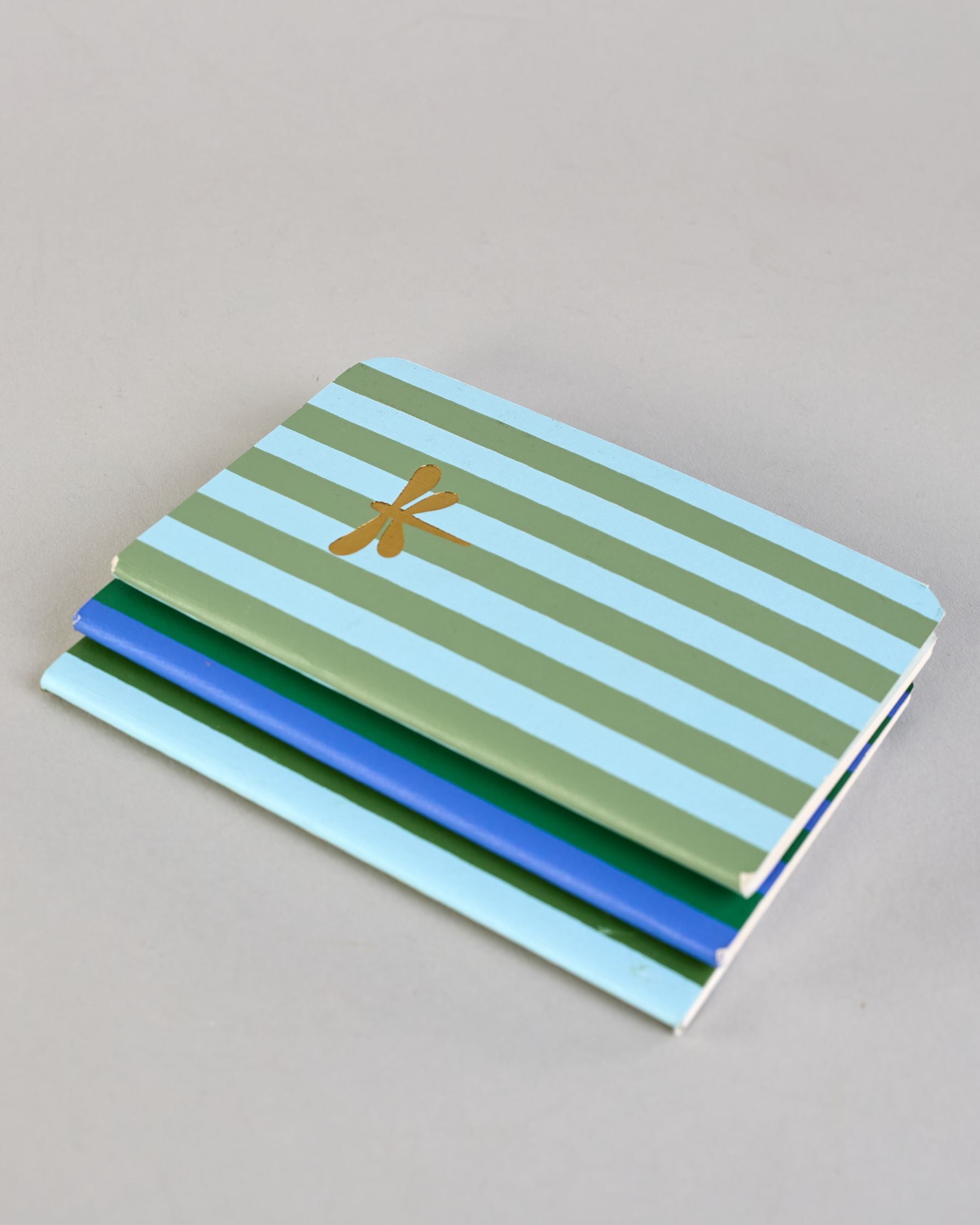 Set aus drei A6-Heften mit recyceltem Baumwolleinband. Die Hefte sind mit senkrechten Streifen in Blau- und Grüntönen gestaltet und zeigen auf dem Cover jeweils ein goldenes Motiv: ein Herz, ein Kleeblatt und eine Libelle. 30 glatte Seiten innen. #11
