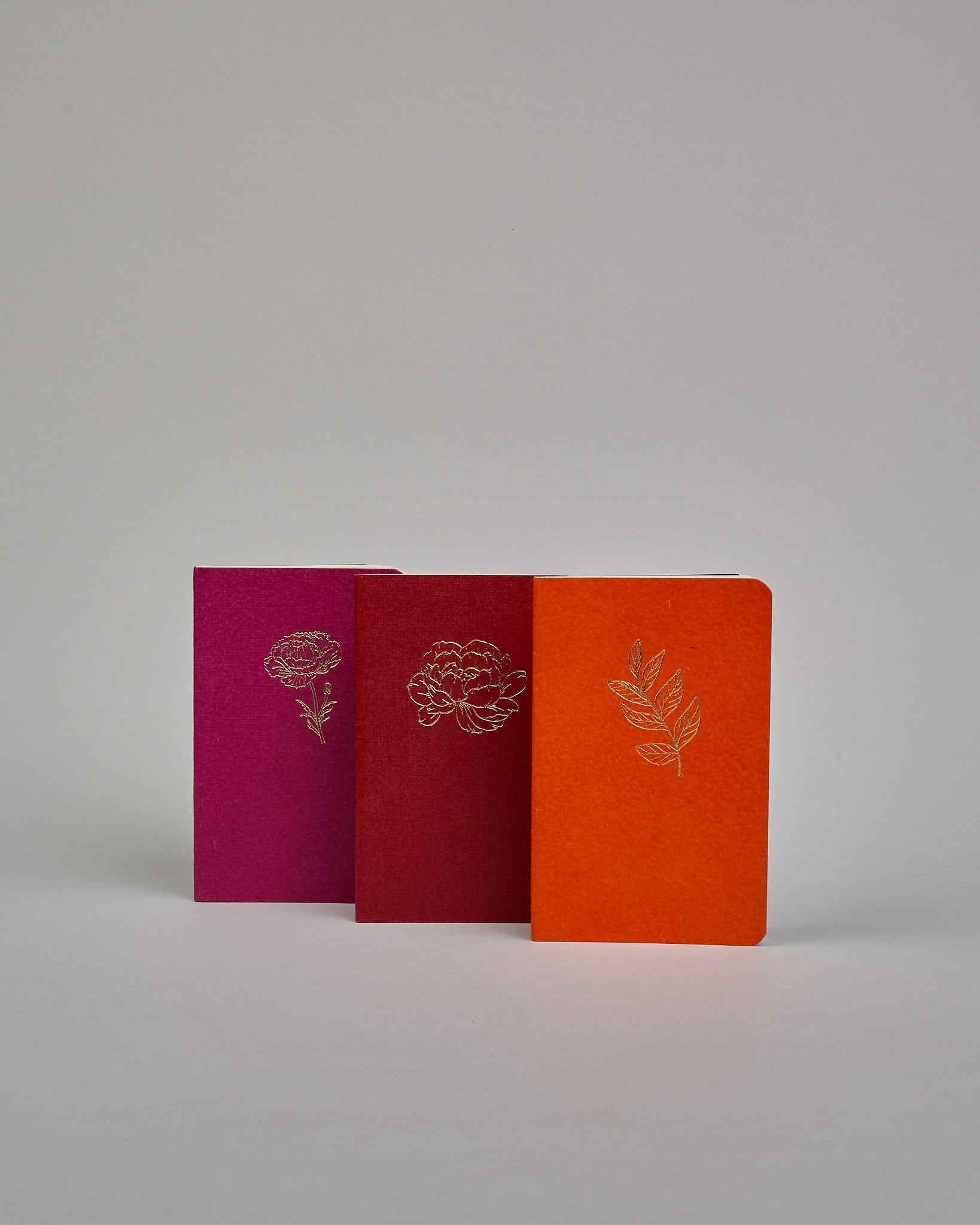 Set bestehend aus drei Heften in magenta, dunkelrot und orange. Jedes hat eine andere Prägung oben Mittig in gold. Das magentafarbene eine Pfingstrose, das dunkelrote eine Rose, das orangene einen Zweig mit Blättern. Innen sind sie glatt. #2