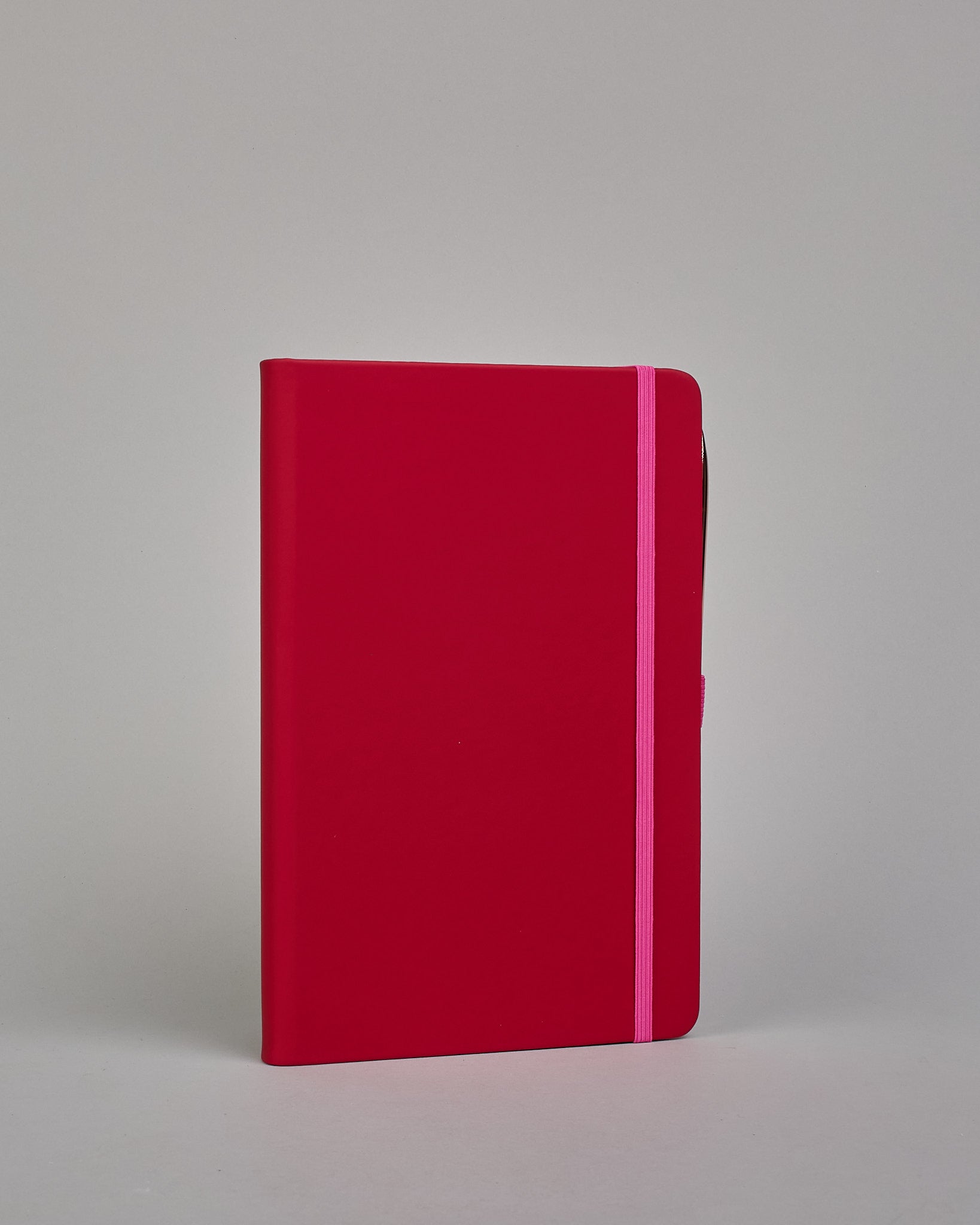 Dunkelrotes Notizbuch mit Soft-Touch Einband, pinkem Gummi zum Verschließen und Lasche aus dem selben Gummi zum Einstecken von einem Stift auf der rechten Seite. Innen finden sich 190 gepunktete Seiten mit Seitennummerierung.