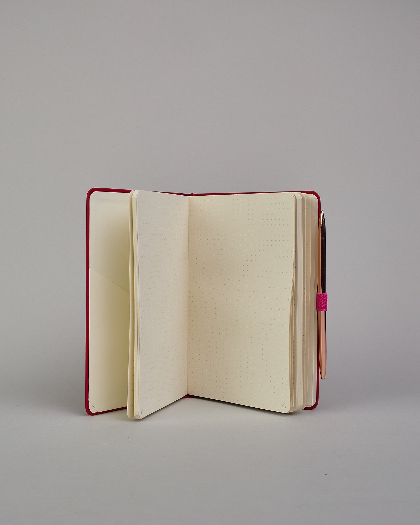 Dunkelrotes Notizbuch mit Soft-Touch Einband, pinkem Gummi zum Verschließen und Lasche aus dem selben Gummi zum Einstecken von einem Stift auf der rechten Seite. Innen finden sich 190 gepunktete Seiten mit Seitennummerierung.