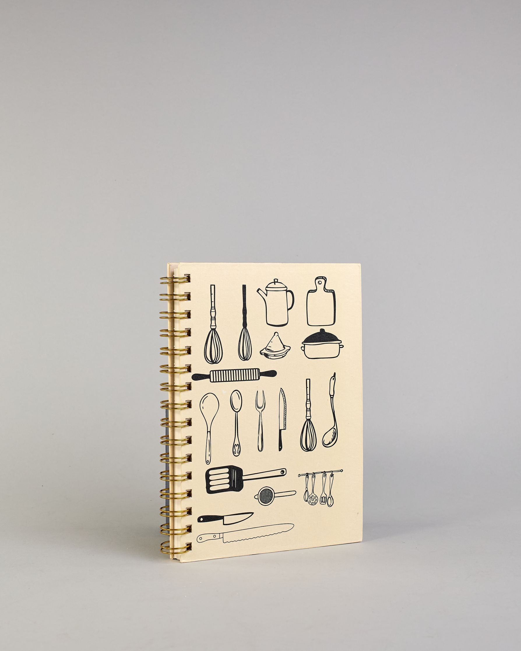 Spiralgebundenes Rezeptbuch mit goldfarbener Wire-O-Bindung und festem cremefarbenem Baumwolleinband aus recycelter Baumwolle. Der Einband ist mit schwarzen Illustrationen von Küchenutensilien wie Schneebesen, Kochlöffel, Messer, Töpfen, Schneidebrett, Pfanne und Sieb dekoriert. Auf den cremeweißen Innenseiten sind vorgedruckte Linien und Felder für das Eintragen von Zutaten und Zubereitungsschritten vorgesehen, jeweils mit klar abgetrennten Bereichen. #2