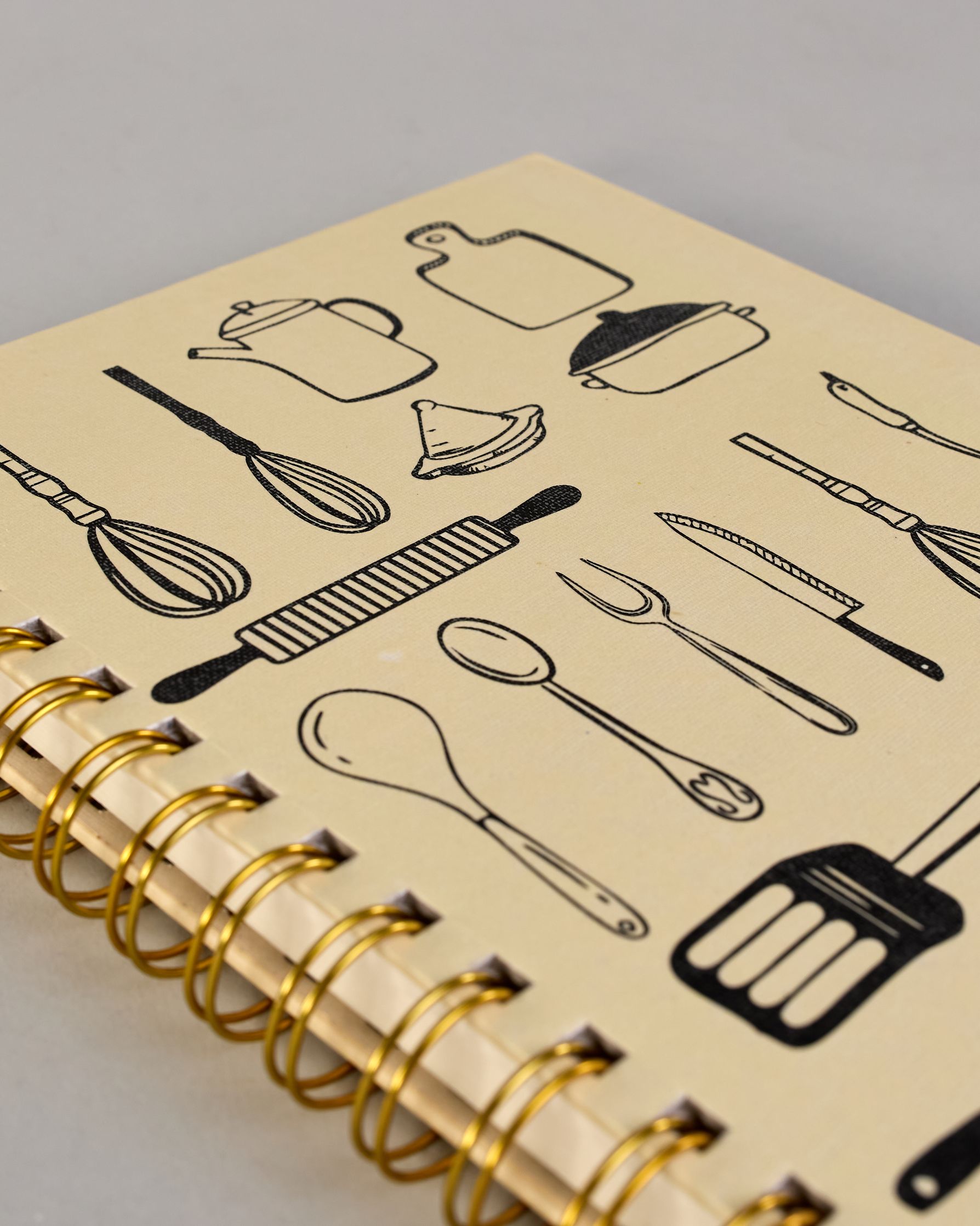 Spiralgebundenes Rezeptbuch mit goldfarbener Wire-O-Bindung und festem cremefarbenem Baumwolleinband aus recycelter Baumwolle. Der Einband ist mit schwarzen Illustrationen von Küchenutensilien wie Schneebesen, Kochlöffel, Messer, Töpfen, Schneidebrett, Pfanne und Sieb dekoriert. Auf den cremeweißen Innenseiten sind vorgedruckte Linien und Felder für das Eintragen von Zutaten und Zubereitungsschritten vorgesehen, jeweils mit klar abgetrennten Bereichen. #4