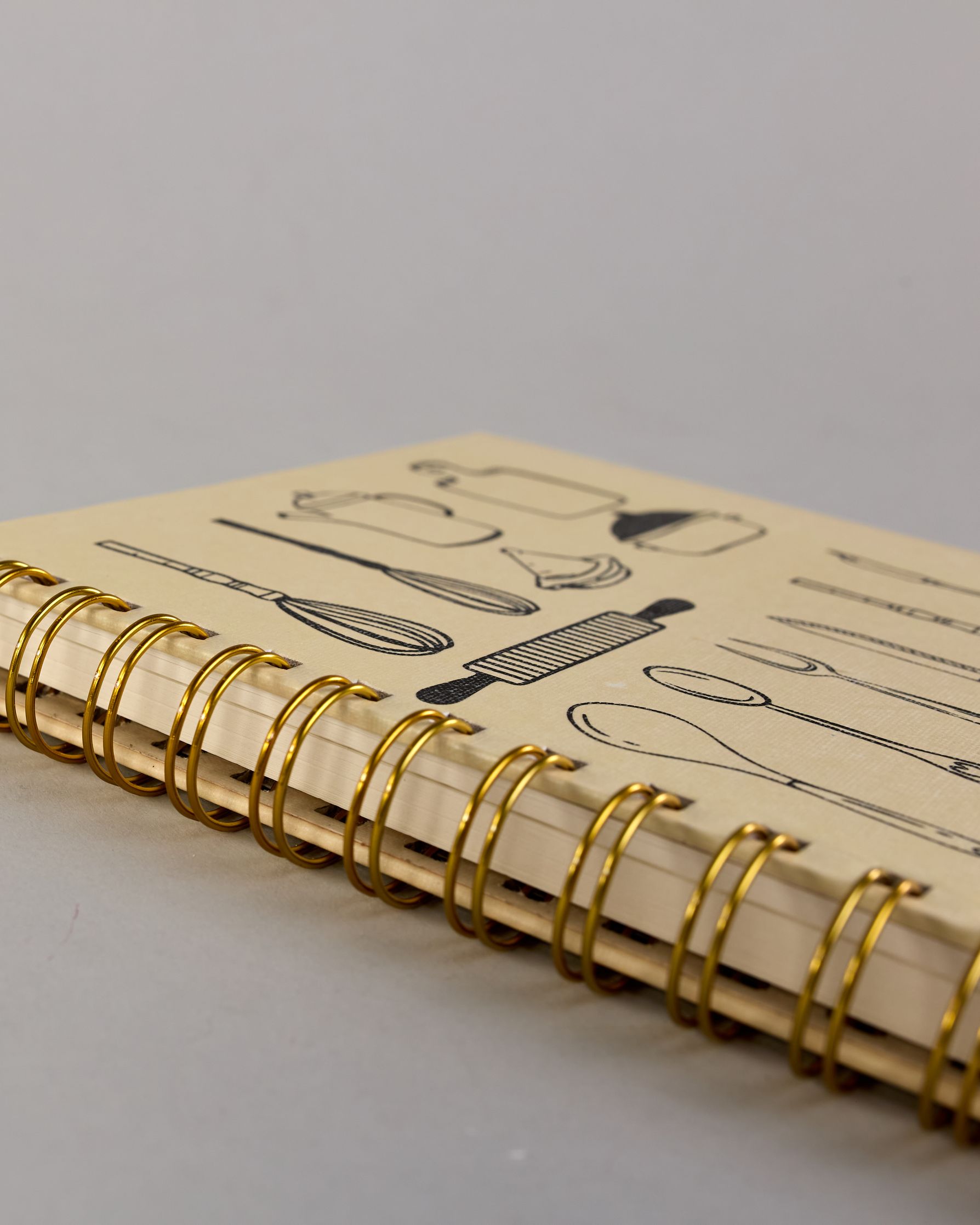 Spiralgebundenes Rezeptbuch mit goldfarbener Wire-O-Bindung und festem cremefarbenem Baumwolleinband aus recycelter Baumwolle. Der Einband ist mit schwarzen Illustrationen von Küchenutensilien wie Schneebesen, Kochlöffel, Messer, Töpfen, Schneidebrett, Pfanne und Sieb dekoriert. Auf den cremeweißen Innenseiten sind vorgedruckte Linien und Felder für das Eintragen von Zutaten und Zubereitungsschritten vorgesehen, jeweils mit klar abgetrennten Bereichen. #5
