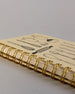 Spiralgebundenes Rezeptbuch mit goldfarbener Wire-O-Bindung und festem cremefarbenem Baumwolleinband aus recycelter Baumwolle. Der Einband ist mit schwarzen Illustrationen von Küchenutensilien wie Schneebesen, Kochlöffel, Messer, Töpfen, Schneidebrett, Pfanne und Sieb dekoriert. Auf den cremeweißen Innenseiten sind vorgedruckte Linien und Felder für das Eintragen von Zutaten und Zubereitungsschritten vorgesehen, jeweils mit klar abgetrennten Bereichen. #5