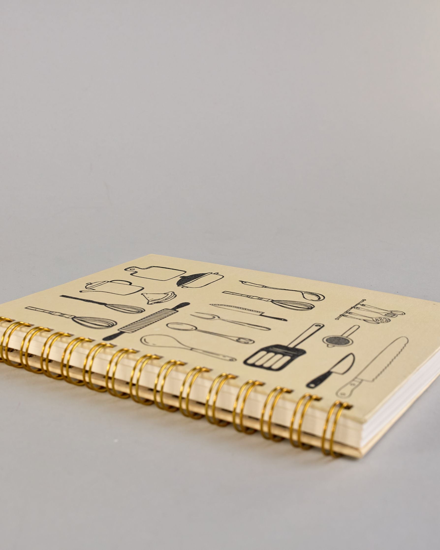 Spiralgebundenes Rezeptbuch mit goldfarbener Wire-O-Bindung und festem cremefarbenem Baumwolleinband aus recycelter Baumwolle. Der Einband ist mit schwarzen Illustrationen von Küchenutensilien wie Schneebesen, Kochlöffel, Messer, Töpfen, Schneidebrett, Pfanne und Sieb dekoriert. Auf den cremeweißen Innenseiten sind vorgedruckte Linien und Felder für das Eintragen von Zutaten und Zubereitungsschritten vorgesehen, jeweils mit klar abgetrennten Bereichen. #7
