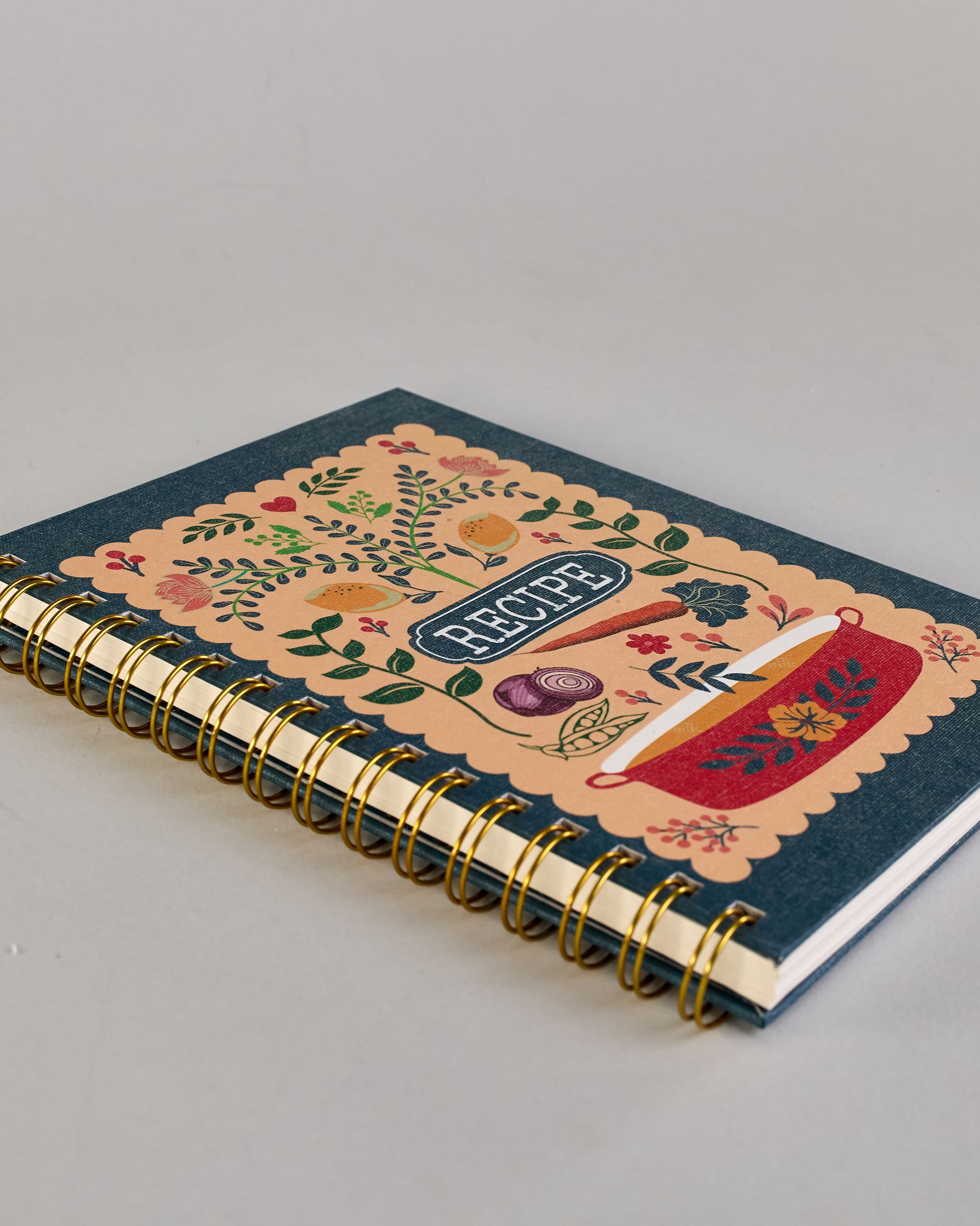 Rezeptbuch mit goldfarbener Spiralbindung und dunkelblauem Einband. Auf der Vorderseite befindet sich ein beigefarbenes Feld mit farbenfrohem, verspieltem Motiv: florale Muster mit Blättern, Blüten, roten Herzen, zwei gelben Vögeln sowie kleinen Illustrationen von Gemüse wie Zwiebel, Karotte und Kohl. Unten ist ein roter Kochtopf mit Blumenornamenten abgebildet. In der Mitte steht das Wort ‚RECIPE‘ in einem dunkelblauen Oval. Auf den cremeweißen Innenseiten sind vorgedruckte Linien und Felder für das Eintr…