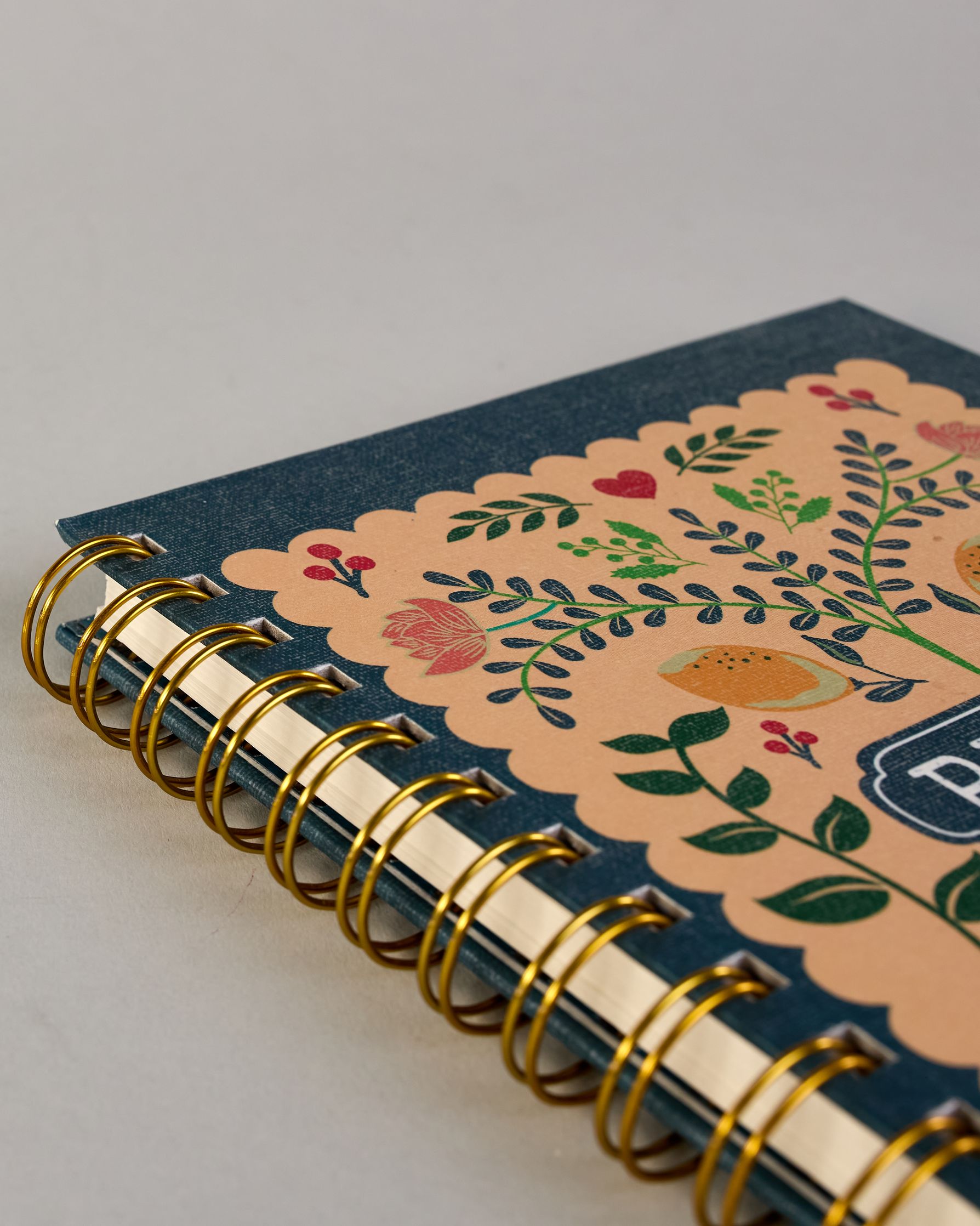 Rezeptbuch mit goldfarbener Spiralbindung und dunkelblauem Einband. Auf der Vorderseite befindet sich ein beigefarbenes Feld mit farbenfrohem, verspieltem Motiv: florale Muster mit Blättern, Blüten, roten Herzen, zwei gelben Vögeln sowie kleinen Illustrationen von Gemüse wie Zwiebel, Karotte und Kohl. Unten ist ein roter Kochtopf mit Blumenornamenten abgebildet. In der Mitte steht das Wort ‚RECIPE‘ in einem dunkelblauen Oval. Auf den cremeweißen Innenseiten sind vorgedruckte Linien und Felder für das Eintr…
