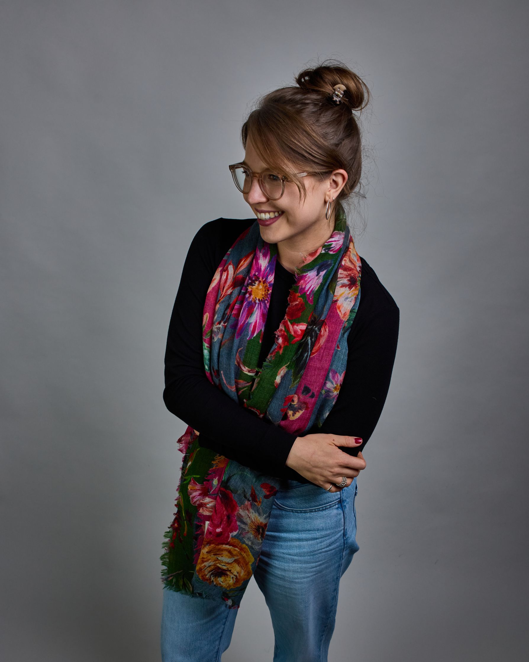 Ein Schal aus 75 % Wolle und 25 % Modal mit den Maßen 70 × 180 cm. Je nach Bild ist er locker um den Nacken gelegt oder einmal gewickelt, um eine lockere Schlaufe um den Hals zu haben, oder zweimal gewickelt, um das Maximum an Wärme zu bekommen. An den Kanten ist er leicht gefranst. Die Hintergrundfarbe des Schals ist blau, und er hat an einer Kante einen magentafarbenen und an der gegenüberliegenden Kante einen grünen Streifen. Auf dem ganzen Schal sind Blumen in Rot-, Orange-, Weiß- und Pinktönen sowie B…