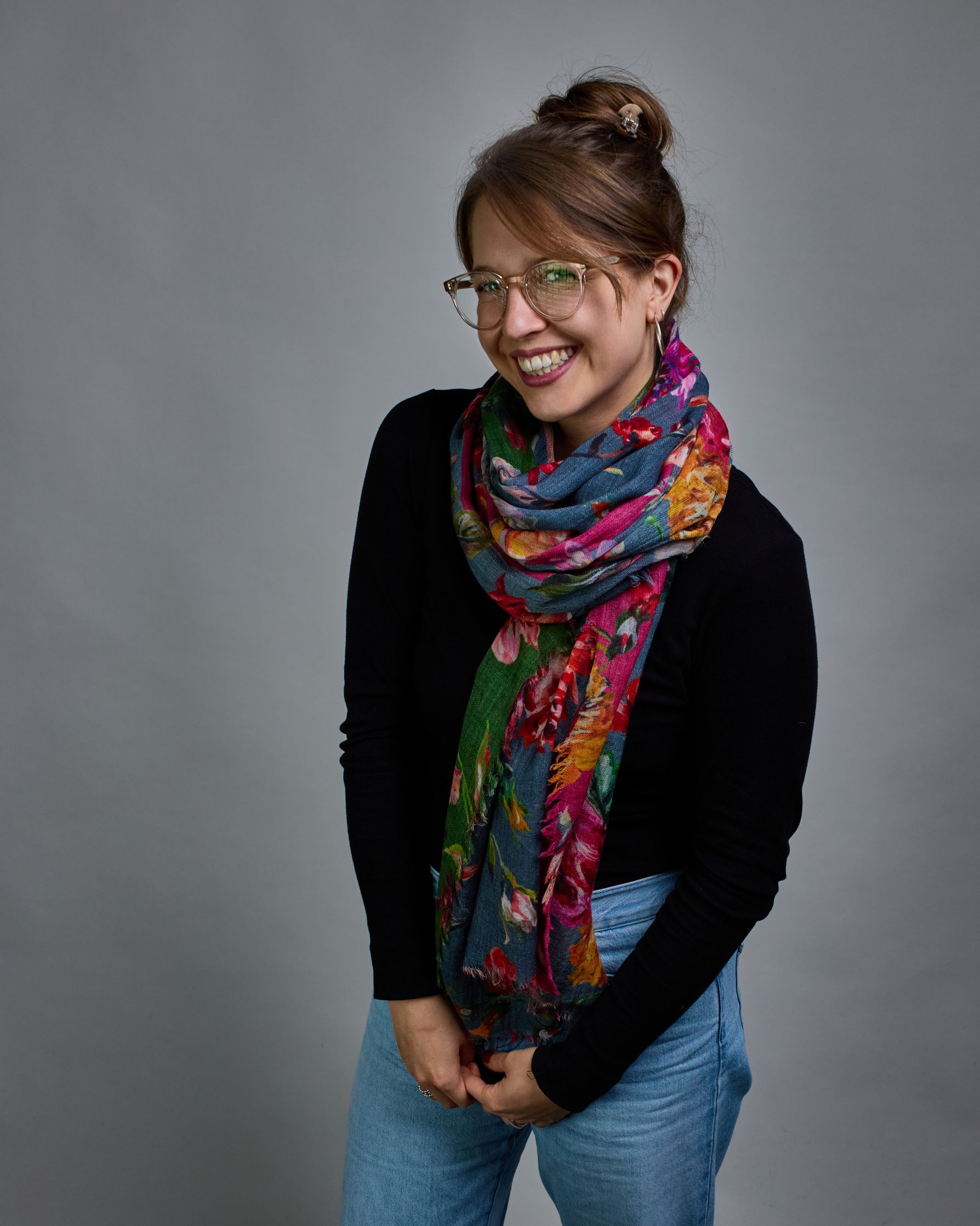 Ein Schal aus 75 % Wolle und 25 % Modal mit den Maßen 70 × 180 cm. Je nach Bild ist er locker um den Nacken gelegt oder einmal gewickelt, um eine lockere Schlaufe um den Hals zu haben, oder zweimal gewickelt, um das Maximum an Wärme zu bekommen. An den Kanten ist er leicht gefranst. Die Hintergrundfarbe des Schals ist blau, und er hat an einer Kante einen magentafarbenen und an der gegenüberliegenden Kante einen grünen Streifen. Auf dem ganzen Schal sind Blumen in Rot-, Orange-, Weiß- und Pinktönen sowie B…