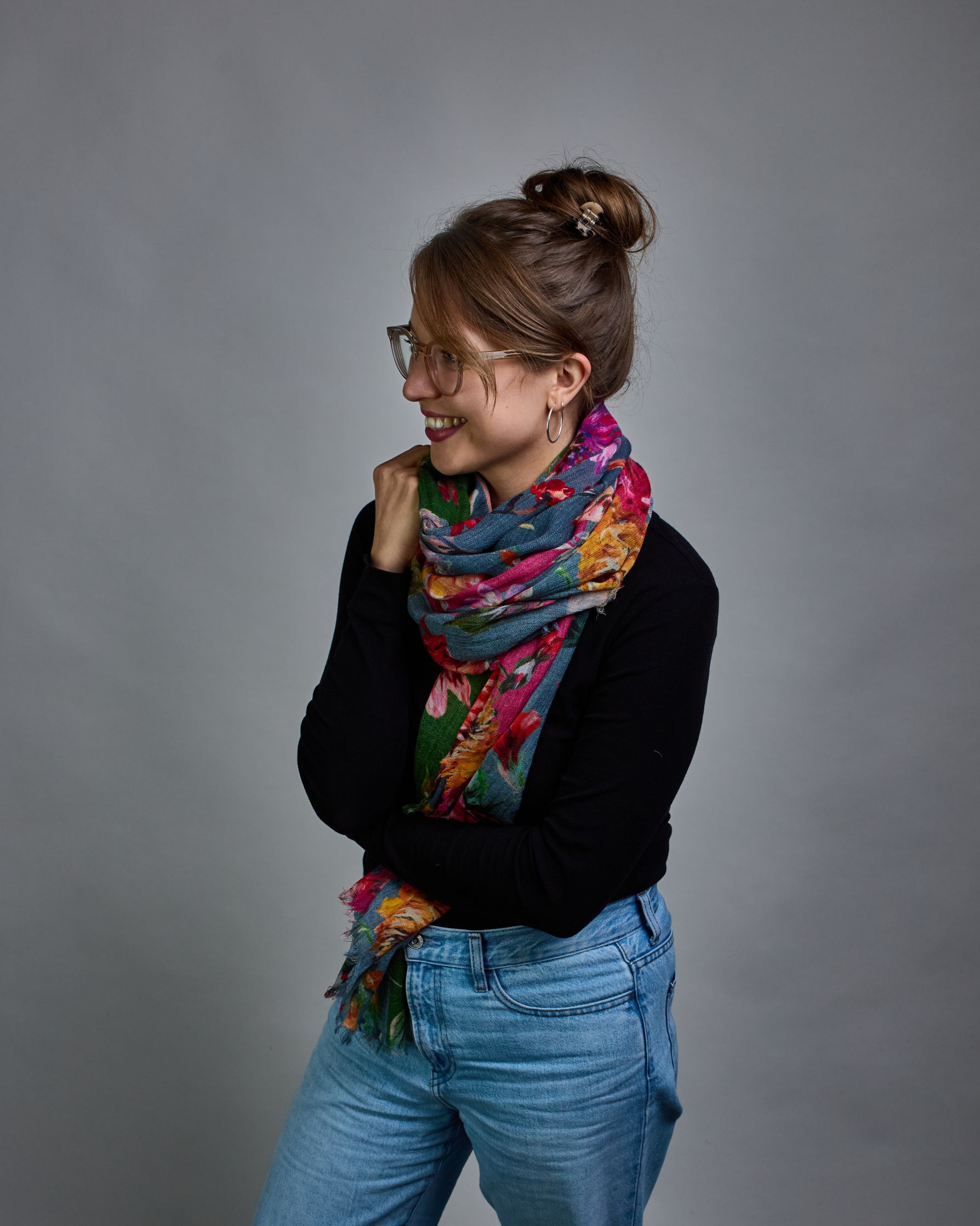 Ein Schal aus 75 % Wolle und 25 % Modal mit den Maßen 70 × 180 cm. Je nach Bild ist er locker um den Nacken gelegt oder einmal gewickelt, um eine lockere Schlaufe um den Hals zu haben, oder zweimal gewickelt, um das Maximum an Wärme zu bekommen. An den Kanten ist er leicht gefranst. Die Hintergrundfarbe des Schals ist blau, und er hat an einer Kante einen magentafarbenen und an der gegenüberliegenden Kante einen grünen Streifen. Auf dem ganzen Schal sind Blumen in Rot-, Orange-, Weiß- und Pinktönen sowie B…