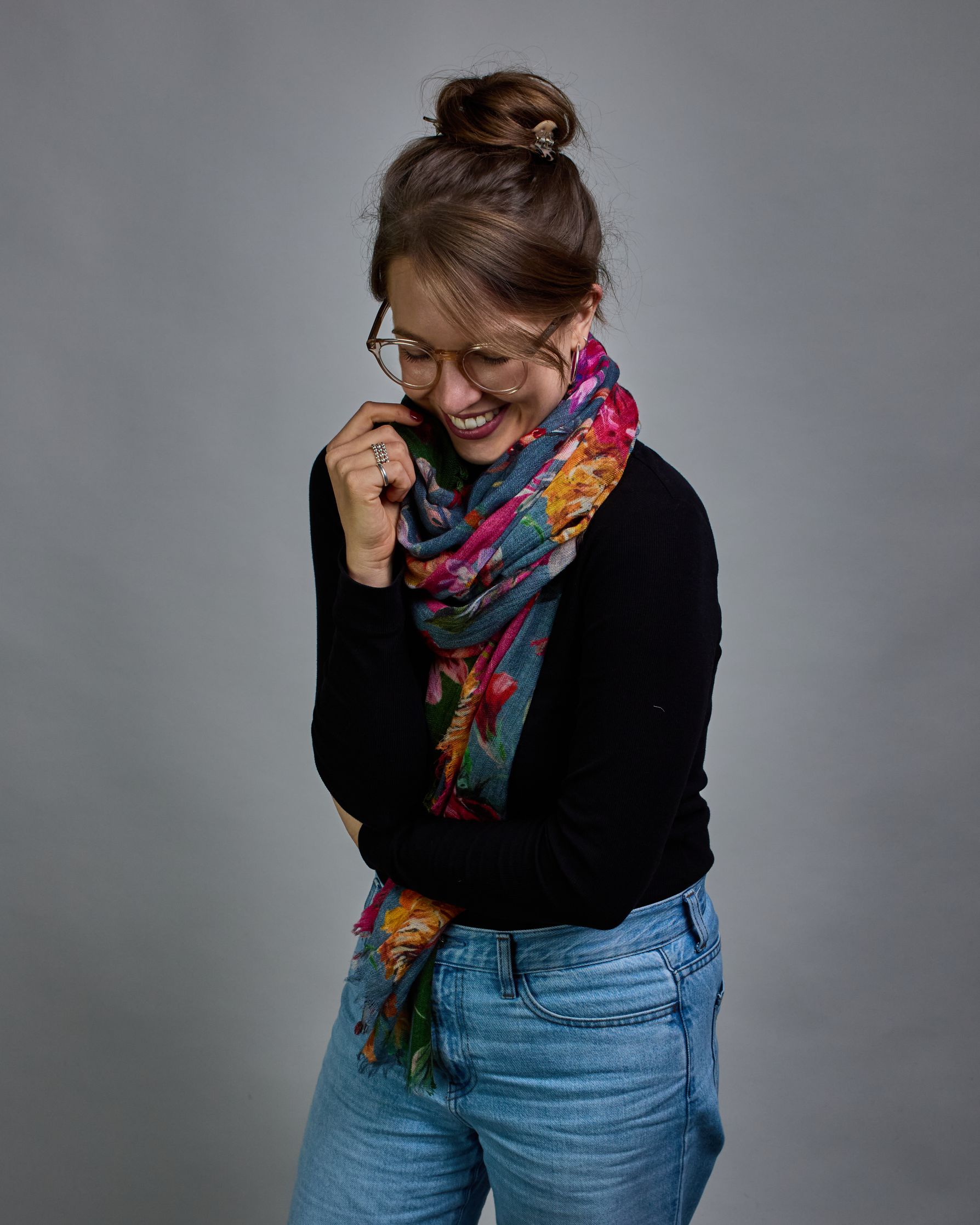 Ein Schal aus 75 % Wolle und 25 % Modal mit den Maßen 70 × 180 cm. Je nach Bild ist er locker um den Nacken gelegt oder einmal gewickelt, um eine lockere Schlaufe um den Hals zu haben, oder zweimal gewickelt, um das Maximum an Wärme zu bekommen. An den Kanten ist er leicht gefranst. Die Hintergrundfarbe des Schals ist blau, und er hat an einer Kante einen magentafarbenen und an der gegenüberliegenden Kante einen grünen Streifen. Auf dem ganzen Schal sind Blumen in Rot-, Orange-, Weiß- und Pinktönen sowie B…