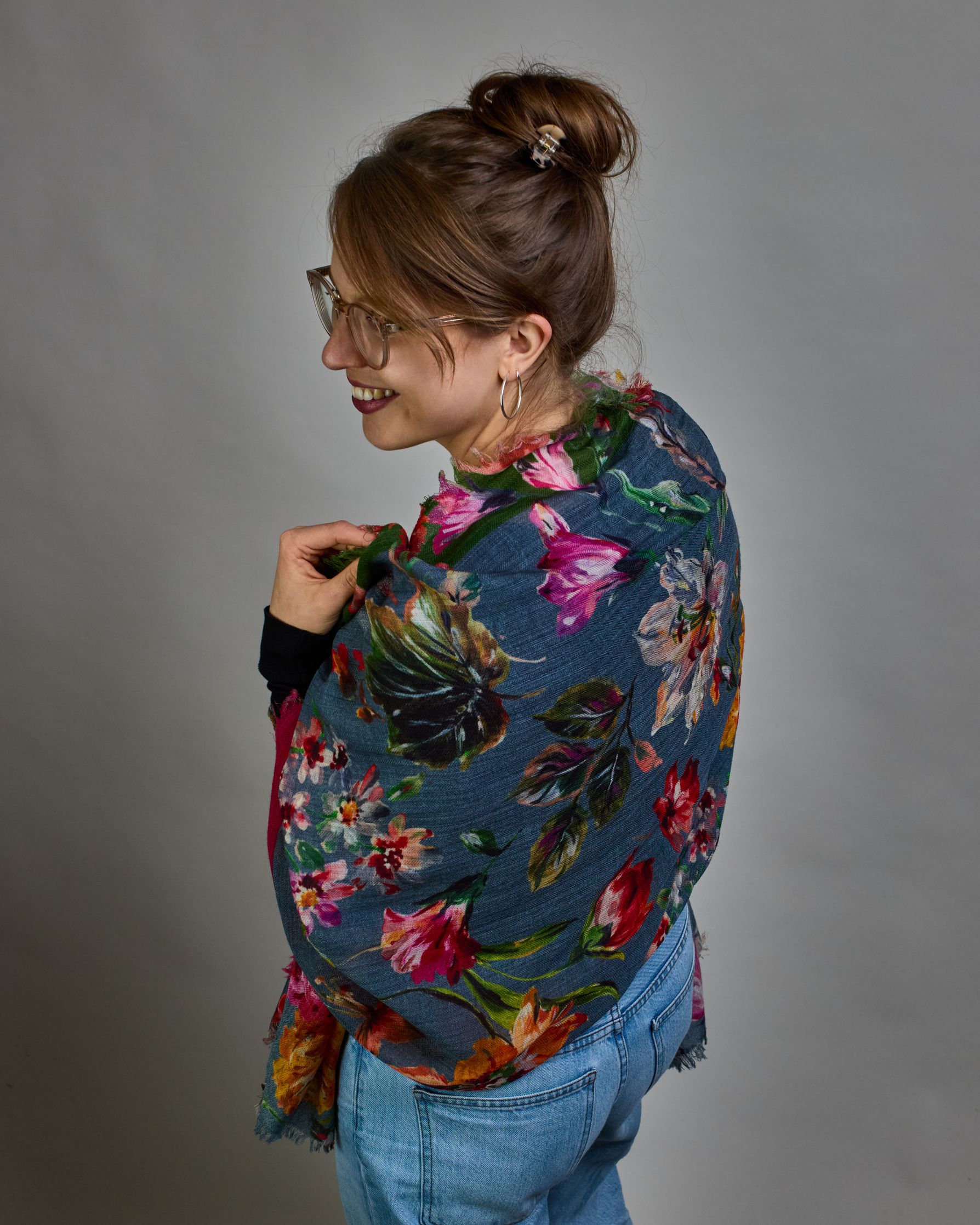 Ein Schal aus 75 % Wolle und 25 % Modal mit den Maßen 70 × 180 cm. Je nach Bild ist er locker um den Nacken gelegt oder einmal gewickelt, um eine lockere Schlaufe um den Hals zu haben, oder zweimal gewickelt, um das Maximum an Wärme zu bekommen. An den Kanten ist er leicht gefranst. Die Hintergrundfarbe des Schals ist blau, und er hat an einer Kante einen magentafarbenen und an der gegenüberliegenden Kante einen grünen Streifen. Auf dem ganzen Schal sind Blumen in Rot-, Orange-, Weiß- und Pinktönen sowie B…