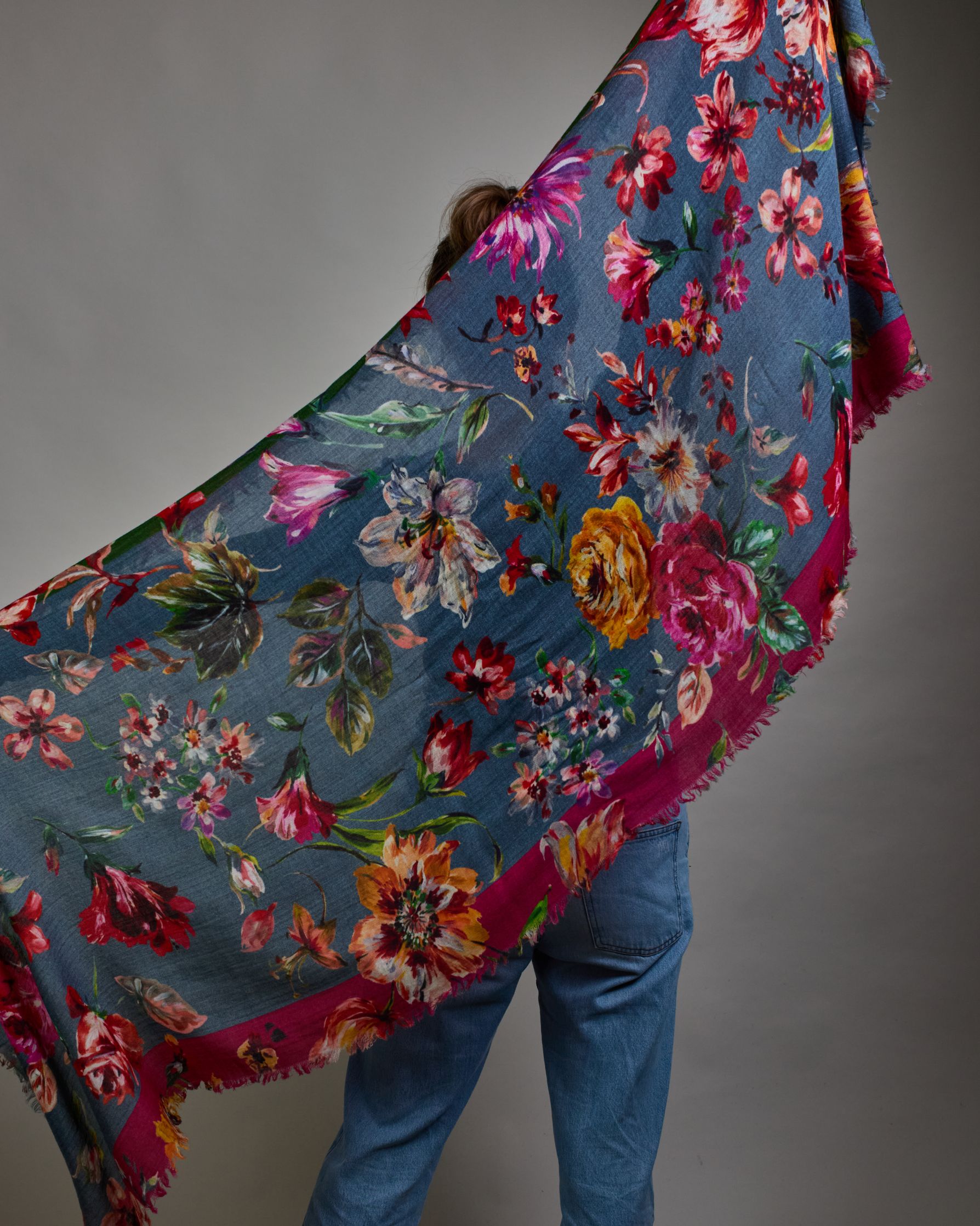 Ein Schal aus 75 % Wolle und 25 % Modal mit den Maßen 70 × 180 cm. Je nach Bild ist er locker um den Nacken gelegt oder einmal gewickelt, um eine lockere Schlaufe um den Hals zu haben, oder zweimal gewickelt, um das Maximum an Wärme zu bekommen. An den Kanten ist er leicht gefranst. Die Hintergrundfarbe des Schals ist blau, und er hat an einer Kante einen magentafarbenen und an der gegenüberliegenden Kante einen grünen Streifen. Auf dem ganzen Schal sind Blumen in Rot-, Orange-, Weiß- und Pinktönen sowie B…