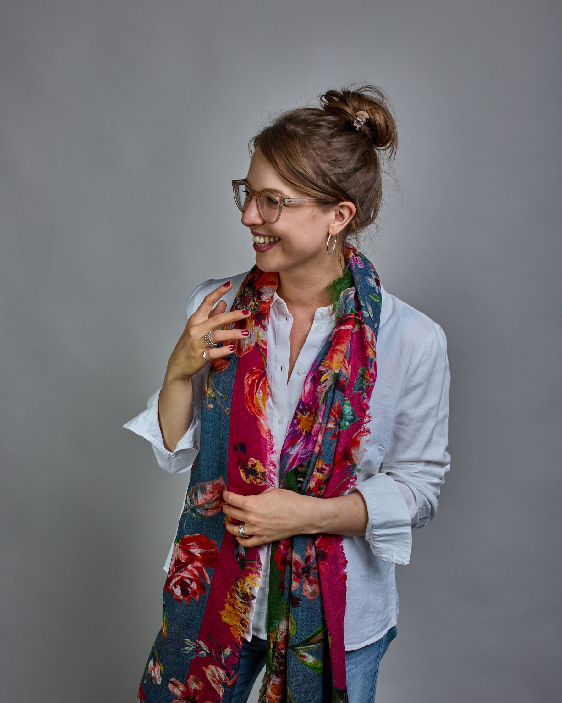 Ein Schal aus 75 % Wolle und 25 % Modal mit den Maßen 70 × 180 cm. Je nach Bild ist er locker um den Nacken gelegt oder einmal gewickelt, um eine lockere Schlaufe um den Hals zu haben, oder zweimal gewickelt, um das Maximum an Wärme zu bekommen. An den Kanten ist er leicht gefranst. Die Hintergrundfarbe des Schals ist blau, und er hat an einer Kante einen magentafarbenen und an der gegenüberliegenden Kante einen grünen Streifen. Auf dem ganzen Schal sind Blumen in Rot-, Orange-, Weiß- und Pinktönen sowie B…