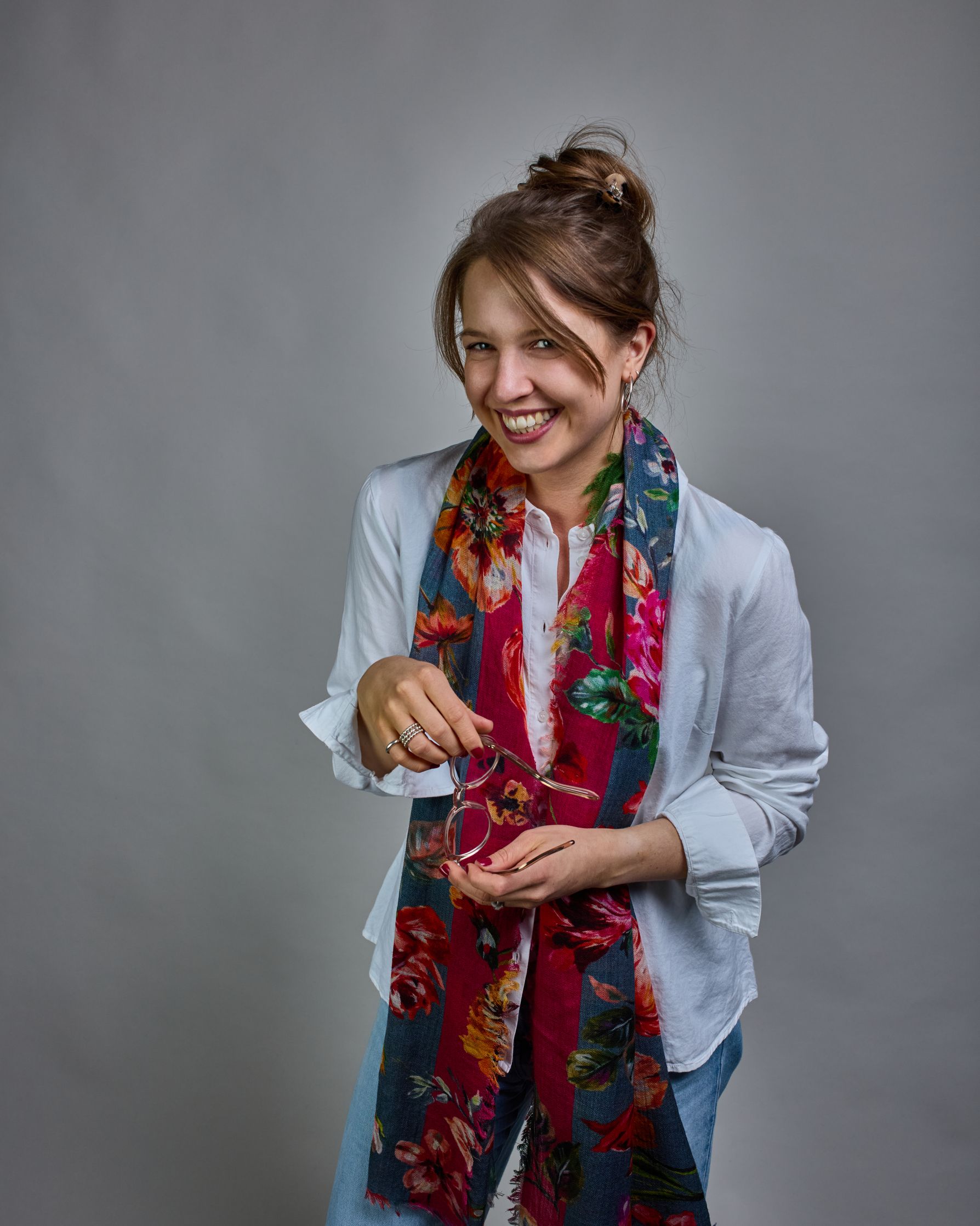 Ein Schal aus 75 % Wolle und 25 % Modal mit den Maßen 70 × 180 cm. Je nach Bild ist er locker um den Nacken gelegt oder einmal gewickelt, um eine lockere Schlaufe um den Hals zu haben, oder zweimal gewickelt, um das Maximum an Wärme zu bekommen. An den Kanten ist er leicht gefranst. Die Hintergrundfarbe des Schals ist blau, und er hat an einer Kante einen magentafarbenen und an der gegenüberliegenden Kante einen grünen Streifen. Auf dem ganzen Schal sind Blumen in Rot-, Orange-, Weiß- und Pinktönen sowie B…