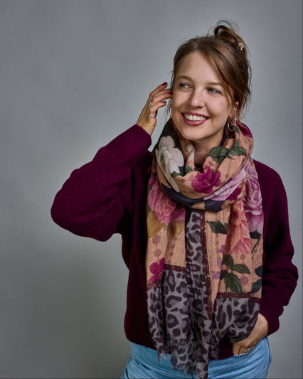 Ein Schal aus 75 % Wolle und 25 % Modal mit den Maßen 70 × 180 cm. Je nach Bild ist er locker um den Nacken gelegt oder einmal gewickelt, um eine lockere Schlaufe um den Hals zu haben, oder zweimal gewickelt, um das Maximum an Wärme zu bekommen. An den Kanten ist er leicht gefranst. Die Hintergrundfarbe des Schals ist ein rosastichiges Beige und an den Kanten ist ein Rahmen in einem Leomuster in verschiedenen Grautönen. Auf dem ganzen Schal sind verschiedene Blüten in unterschiedlichen Rosa-, und Weißtönen…