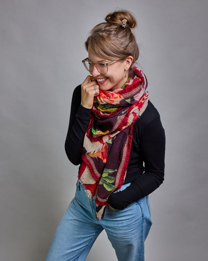 Ein Schal aus 75 % Wolle und 25 % Modal mit den Maßen 70 × 180 cm. Je nach Bild ist er locker um den Nacken gelegt oder einmal gewickelt, um eine lockere Schlaufe um den Hals zu haben, oder zweimal gewickelt, um das Maximum an Wärme zu bekommen. An den Kanten ist er leicht gefranst. Die Hintergrundfarbe des Schals ist schwarz, und er hat an den Kanten einen rot-weiß gemusterten Rahmen. Zwischen diesem Rahmen und dem restlichen Design des Schals ist in einem festen Stoff eine schmale symmetrisch gemusterte…