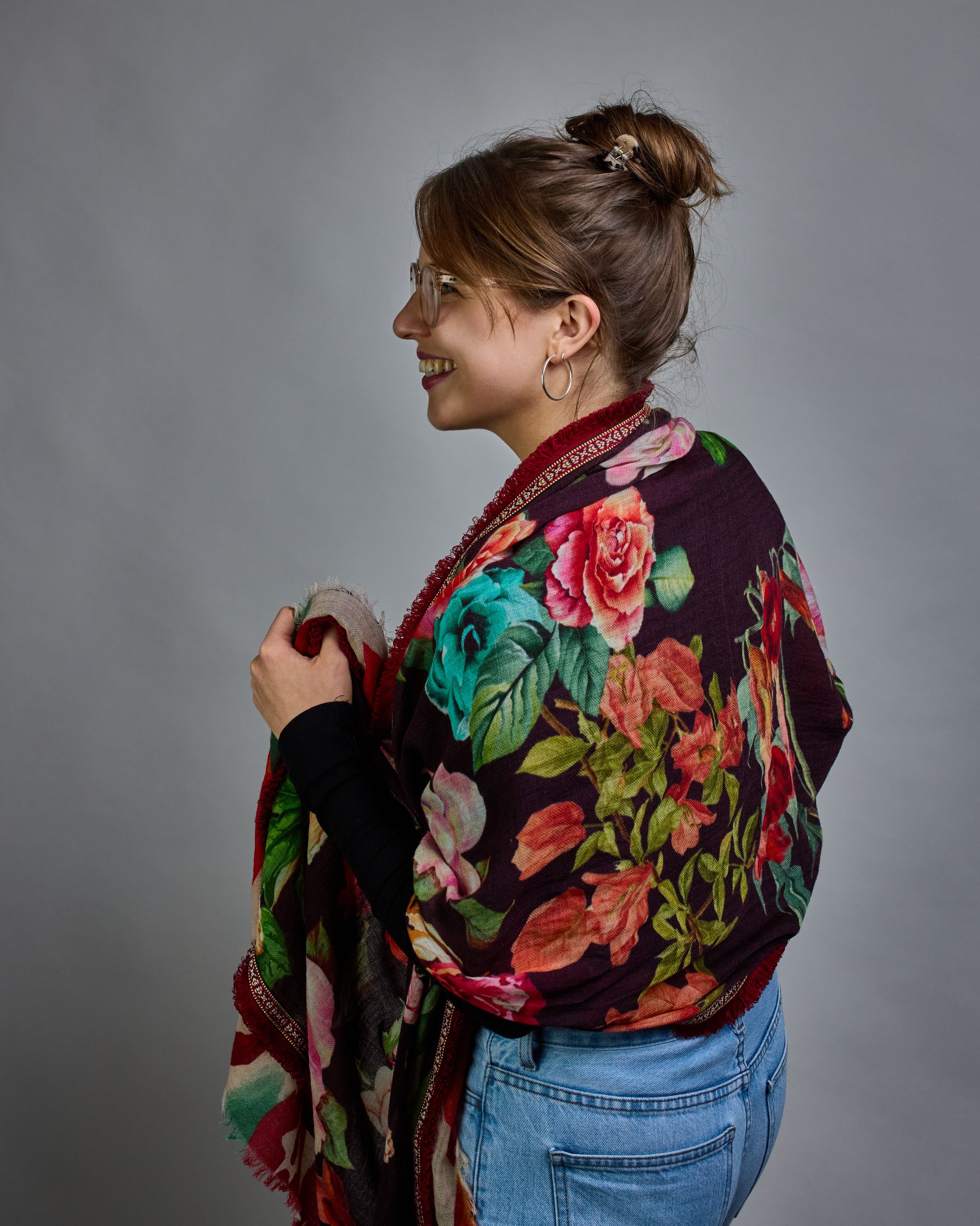 Ein Schal aus 75 % Wolle und 25 % Modal mit den Maßen 70 × 180 cm. Je nach Bild ist er locker um den Nacken gelegt oder einmal gewickelt, um eine lockere Schlaufe um den Hals zu haben, oder zweimal gewickelt, um das Maximum an Wärme zu bekommen. An den Kanten ist er leicht gefranst. Die Hintergrundfarbe des Schals ist schwarz, und er hat an den Kanten einen rot-weiß gemusterten Rahmen. Zwischen diesem Rahmen und dem restlichen Design des Schals ist in einem festen Stoff eine schmale symmetrisch gemusterte…