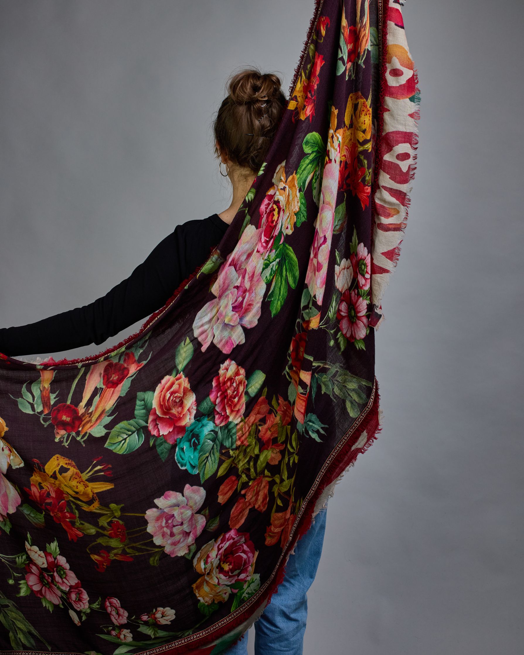 Ein Schal aus 75 % Wolle und 25 % Modal mit den Maßen 70 × 180 cm. Je nach Bild ist er locker um den Nacken gelegt oder einmal gewickelt, um eine lockere Schlaufe um den Hals zu haben, oder zweimal gewickelt, um das Maximum an Wärme zu bekommen. An den Kanten ist er leicht gefranst. Die Hintergrundfarbe des Schals ist schwarz, und er hat an den Kanten einen rot-weiß gemusterten Rahmen. Zwischen diesem Rahmen und dem restlichen Design des Schals ist in einem festen Stoff eine schmale symmetrisch gemusterte…