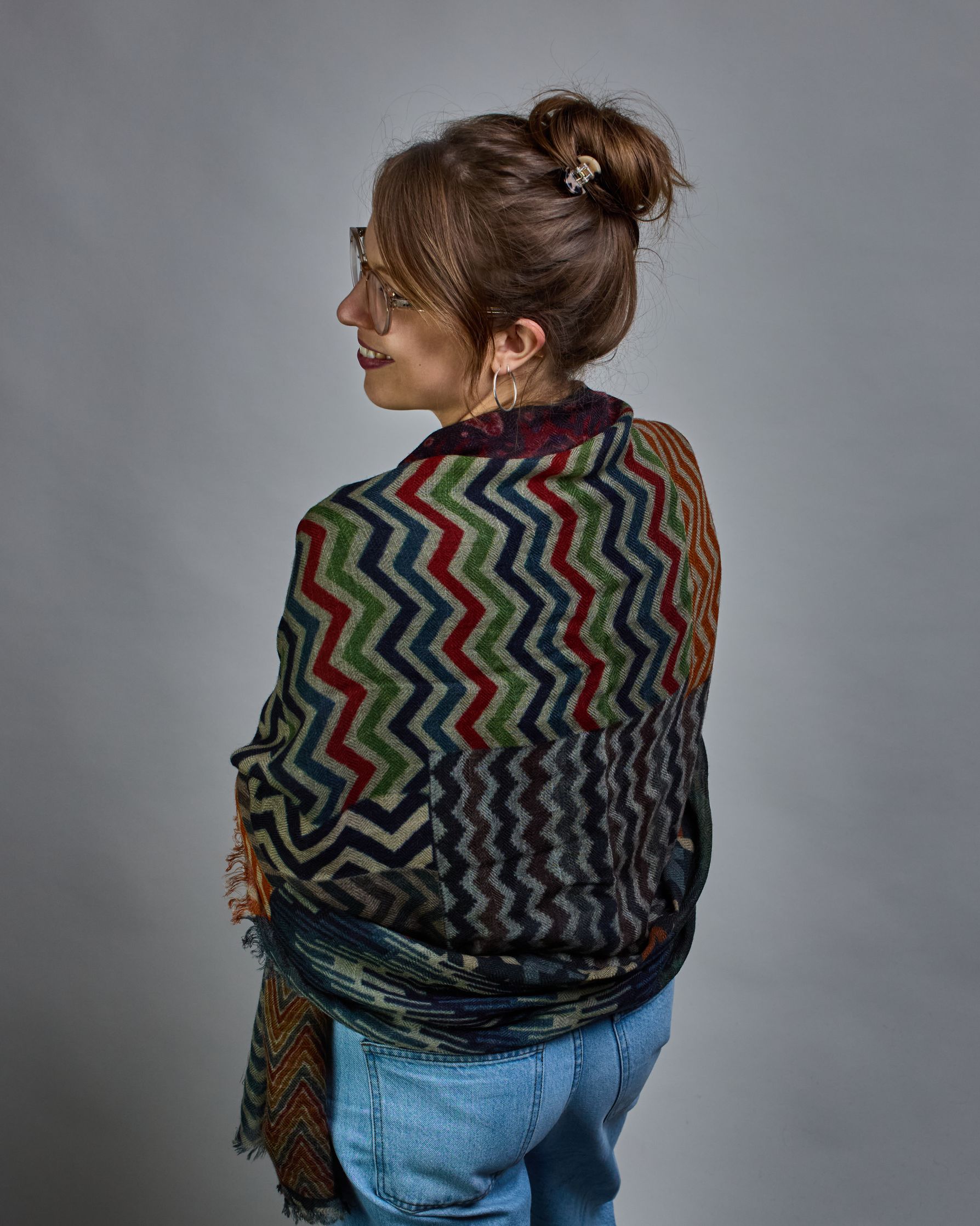 Ein Schal aus 75 % Wolle und 25 % Modal mit den Maßen 100 × 180 cm. Je nach Bild ist er locker um den Nacken gelegt oder einmal gewickelt, um eine lockere Schlaufe um den Hals zu haben, oder zweimal gewickelt, um das Maximum an Wärme zu bekommen. An den Kanten ist er leicht gefranst. Der Schal ist in einem Patchworkstil designt, in welchem jedes Rechteck Zickzackmuster in unterschiedlichen Größen und Farben hat. Die primären Farben sind verschiedene Grautöne, Orange, Rot, Grün, Blautöne, Türkis und Beige.…