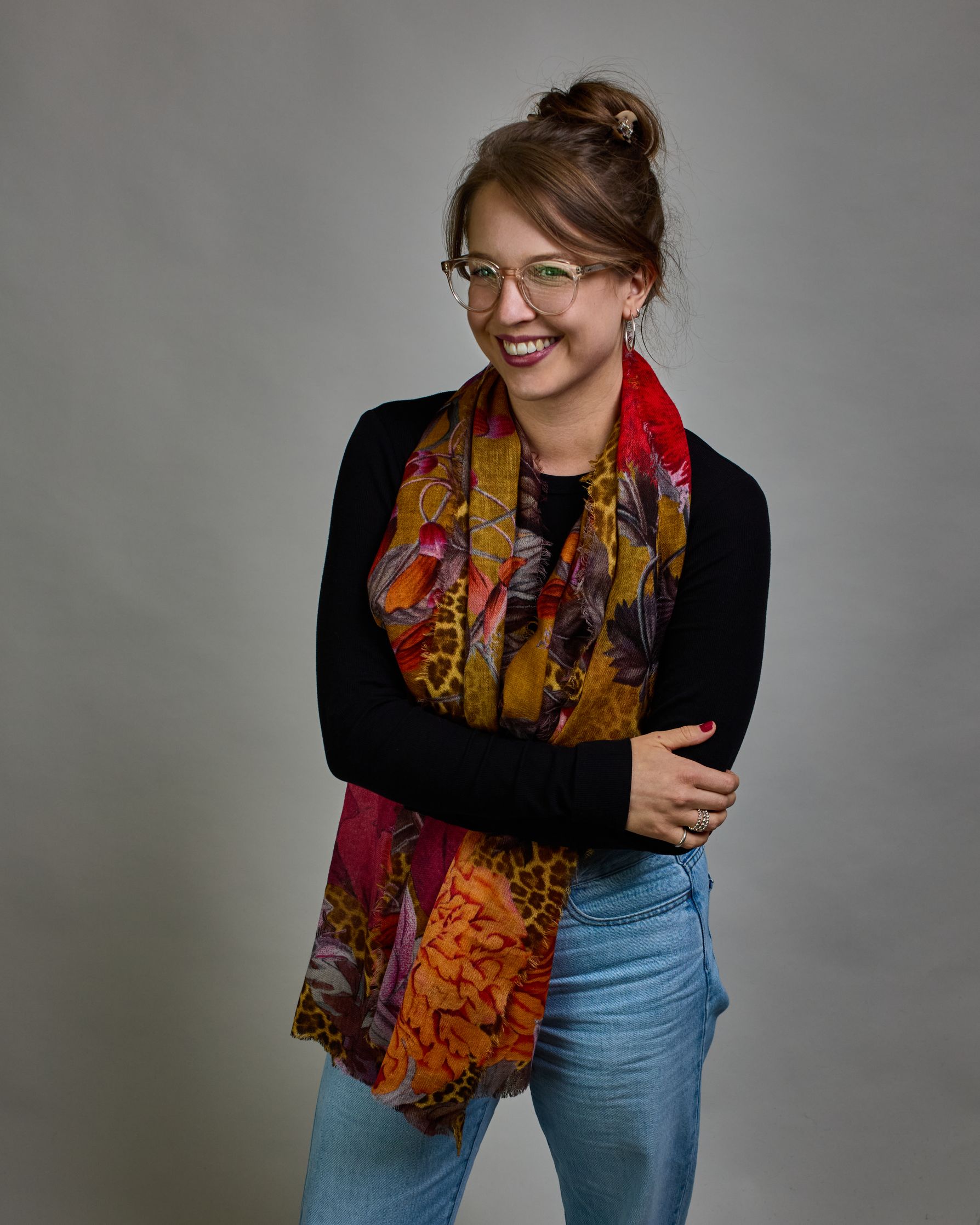 Ein Schal aus 75 % Wolle und 25 % Modal mit den Maßen 70 × 180 cm. Je nach Bild ist er locker um den Nacken gelegt oder einmal gewickelt, um eine lockere Schlaufe um den Hals zu haben, oder zweimal gewickelt, um das Maximum an Wärme zu bekommen. An den Kanten ist er leicht gefranst. Die Hintergrundfarbe des Schals ist Okker und hier und da ist ein Leomuster in Brauntönen zu sehen. Auf dem ganzen Schal sind Blumen in Rot-, Orange-, Weiß- und Pinktönen sowie Blätter beziehungsweise Blumenstängel in verschied…