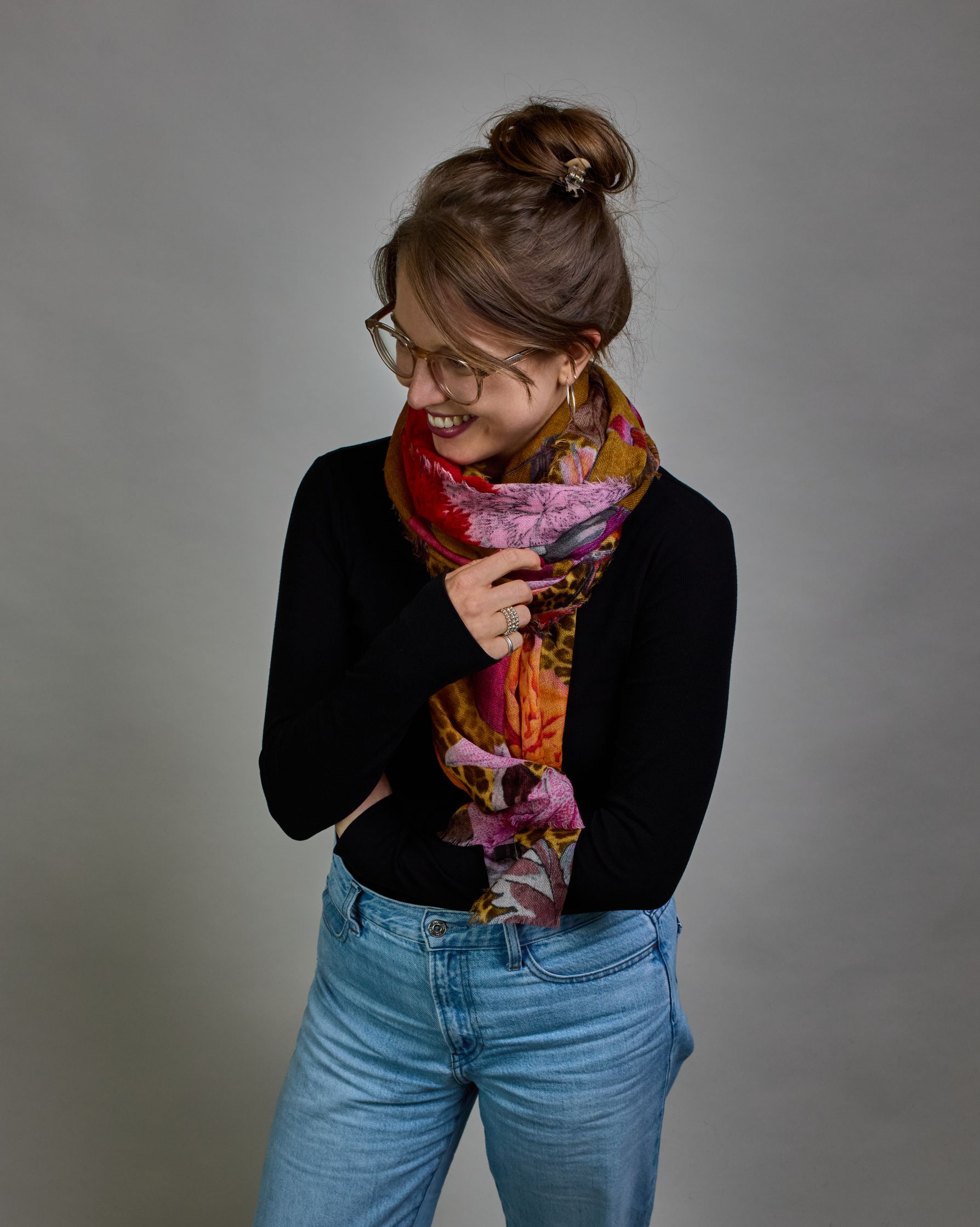 Ein Schal aus 75 % Wolle und 25 % Modal mit den Maßen 70 × 180 cm. Je nach Bild ist er locker um den Nacken gelegt oder einmal gewickelt, um eine lockere Schlaufe um den Hals zu haben, oder zweimal gewickelt, um das Maximum an Wärme zu bekommen. An den Kanten ist er leicht gefranst. Die Hintergrundfarbe des Schals ist Okker und hier und da ist ein Leomuster in Brauntönen zu sehen. Auf dem ganzen Schal sind Blumen in Rot-, Orange-, Weiß- und Pinktönen sowie Blätter beziehungsweise Blumenstängel in verschied…