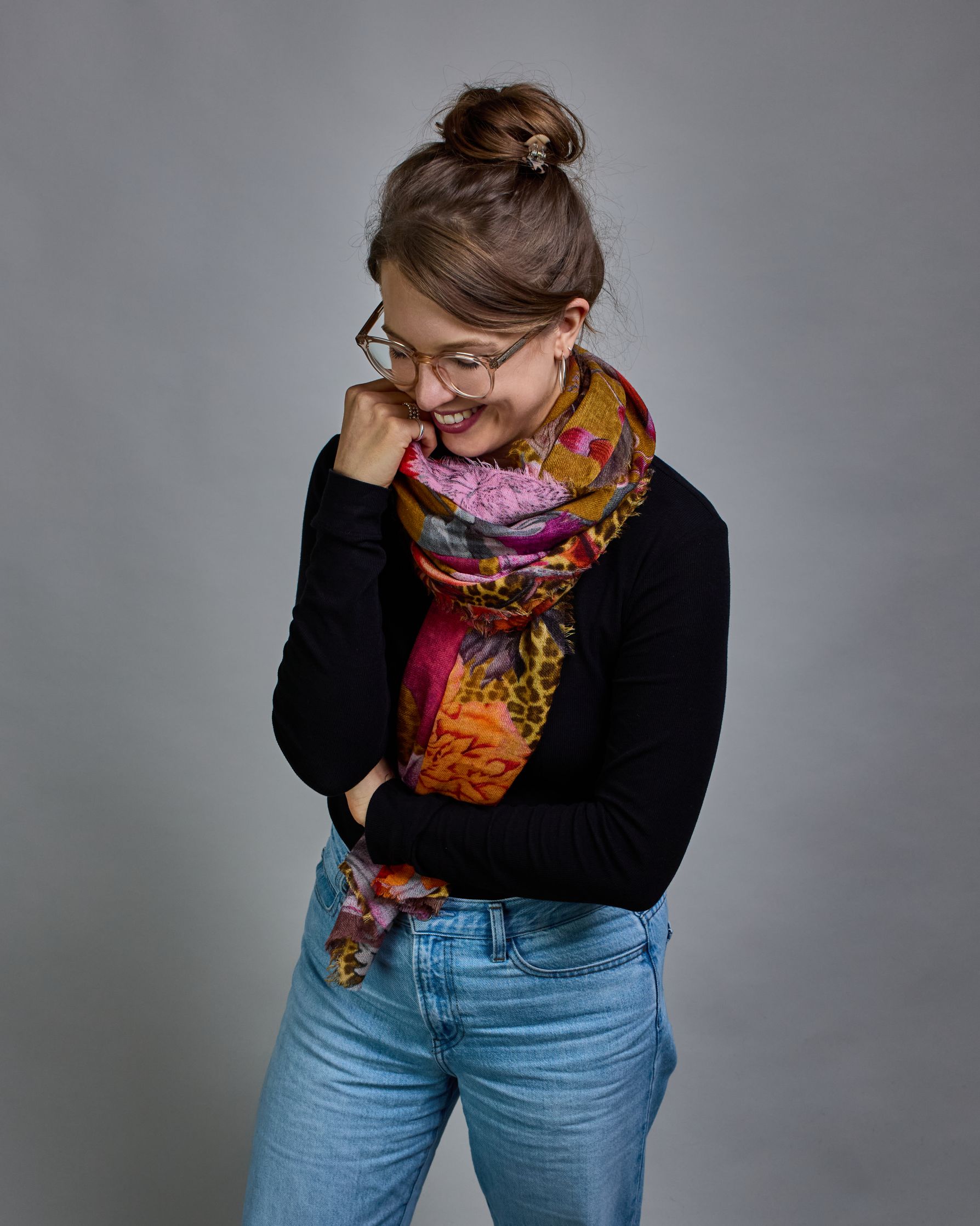 Ein Schal aus 75 % Wolle und 25 % Modal mit den Maßen 70 × 180 cm. Je nach Bild ist er locker um den Nacken gelegt oder einmal gewickelt, um eine lockere Schlaufe um den Hals zu haben, oder zweimal gewickelt, um das Maximum an Wärme zu bekommen. An den Kanten ist er leicht gefranst. Die Hintergrundfarbe des Schals ist Okker und hier und da ist ein Leomuster in Brauntönen zu sehen. Auf dem ganzen Schal sind Blumen in Rot-, Orange-, Weiß- und Pinktönen sowie Blätter beziehungsweise Blumenstängel in verschied…