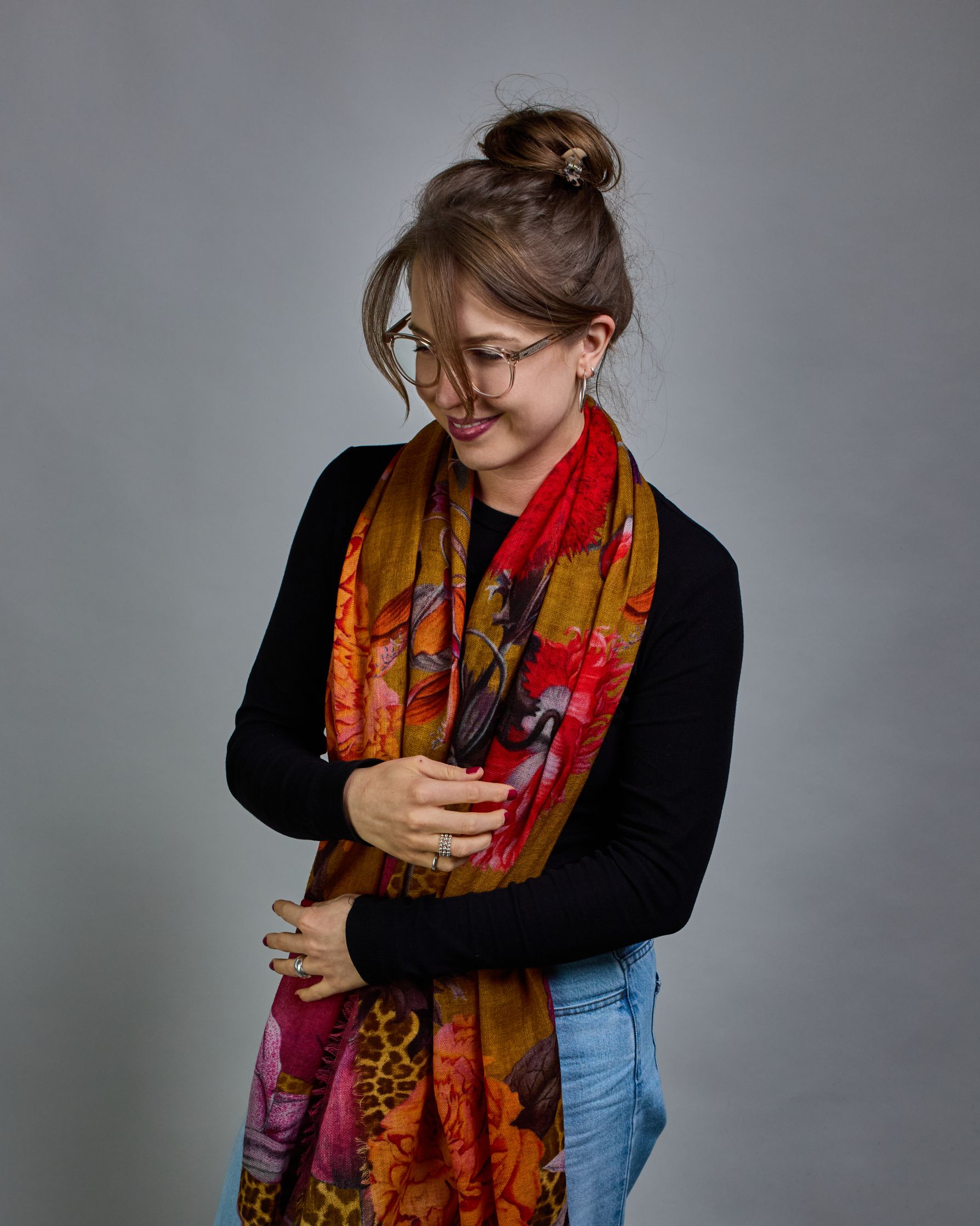 Ein Schal aus 75 % Wolle und 25 % Modal mit den Maßen 70 × 180 cm. Je nach Bild ist er locker um den Nacken gelegt oder einmal gewickelt, um eine lockere Schlaufe um den Hals zu haben, oder zweimal gewickelt, um das Maximum an Wärme zu bekommen. An den Kanten ist er leicht gefranst. Die Hintergrundfarbe des Schals ist Okker und hier und da ist ein Leomuster in Brauntönen zu sehen. Auf dem ganzen Schal sind Blumen in Rot-, Orange-, Weiß- und Pinktönen sowie Blätter beziehungsweise Blumenstängel in verschied…