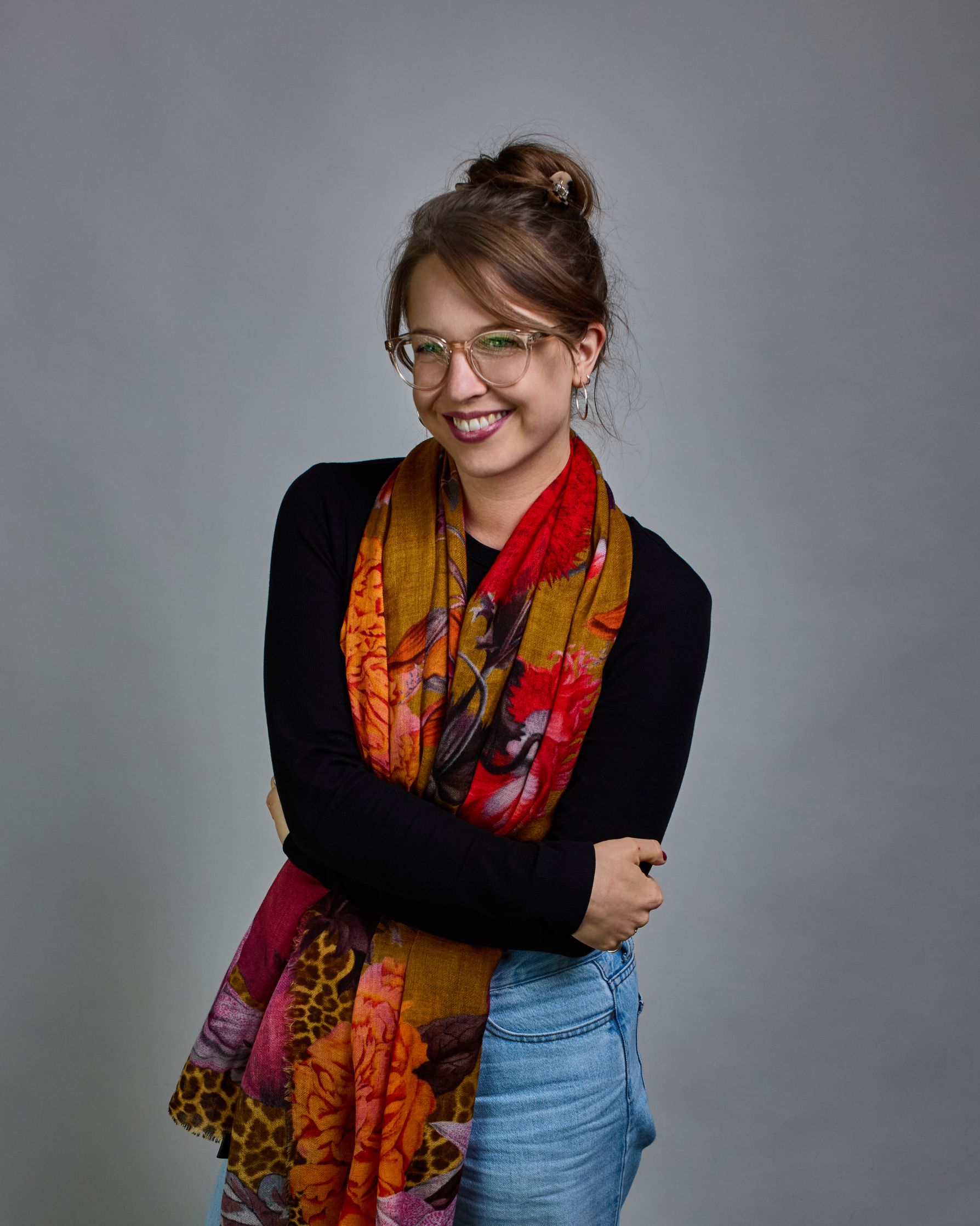 Ein Schal aus 75 % Wolle und 25 % Modal mit den Maßen 70 × 180 cm. Je nach Bild ist er locker um den Nacken gelegt oder einmal gewickelt, um eine lockere Schlaufe um den Hals zu haben, oder zweimal gewickelt, um das Maximum an Wärme zu bekommen. An den Kanten ist er leicht gefranst. Die Hintergrundfarbe des Schals ist Okker und hier und da ist ein Leomuster in Brauntönen zu sehen. Auf dem ganzen Schal sind Blumen in Rot-, Orange-, Weiß- und Pinktönen sowie Blätter beziehungsweise Blumenstängel in verschied…