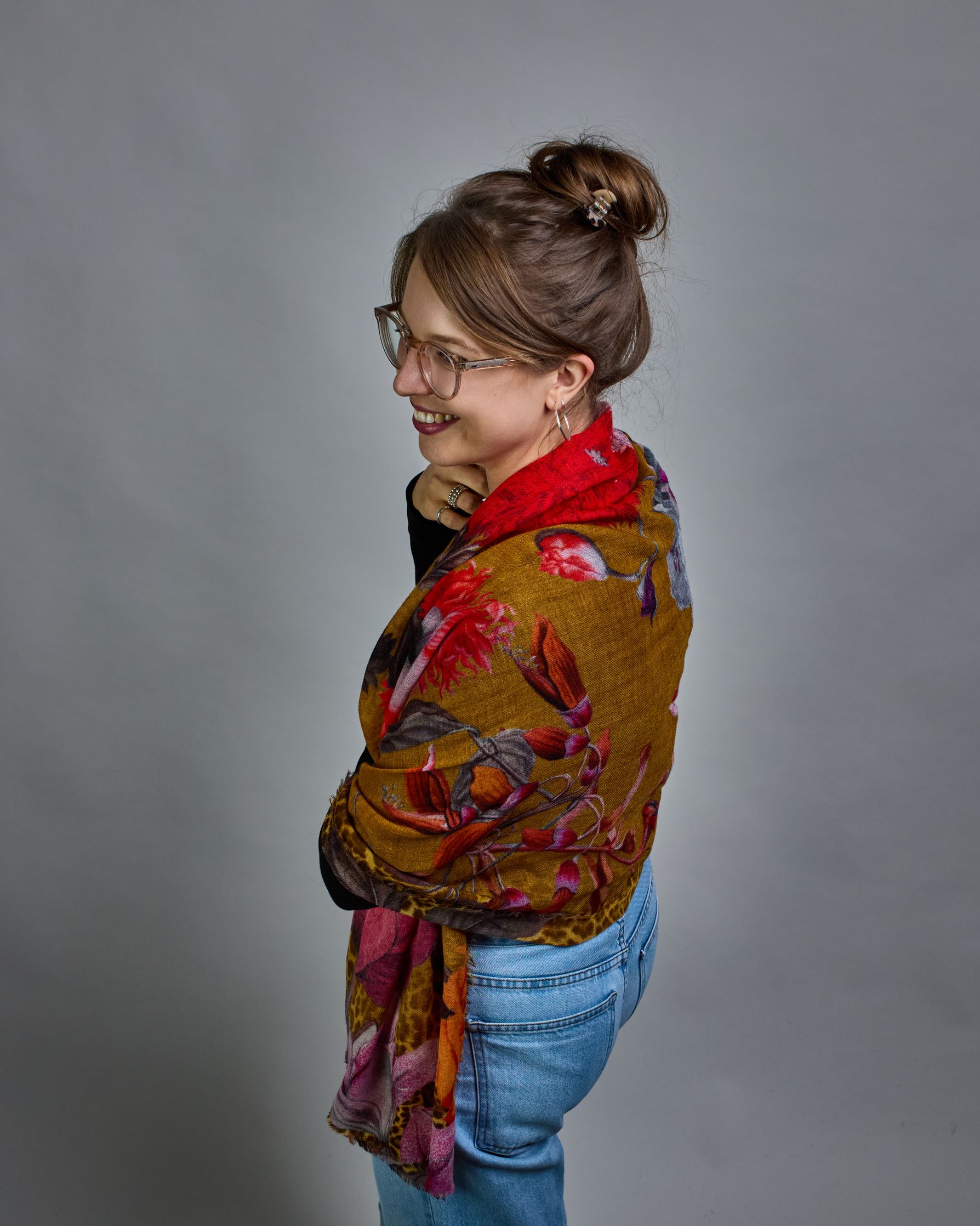 Ein Schal aus 75 % Wolle und 25 % Modal mit den Maßen 70 × 180 cm. Je nach Bild ist er locker um den Nacken gelegt oder einmal gewickelt, um eine lockere Schlaufe um den Hals zu haben, oder zweimal gewickelt, um das Maximum an Wärme zu bekommen. An den Kanten ist er leicht gefranst. Die Hintergrundfarbe des Schals ist Okker und hier und da ist ein Leomuster in Brauntönen zu sehen. Auf dem ganzen Schal sind Blumen in Rot-, Orange-, Weiß- und Pinktönen sowie Blätter beziehungsweise Blumenstängel in verschied…