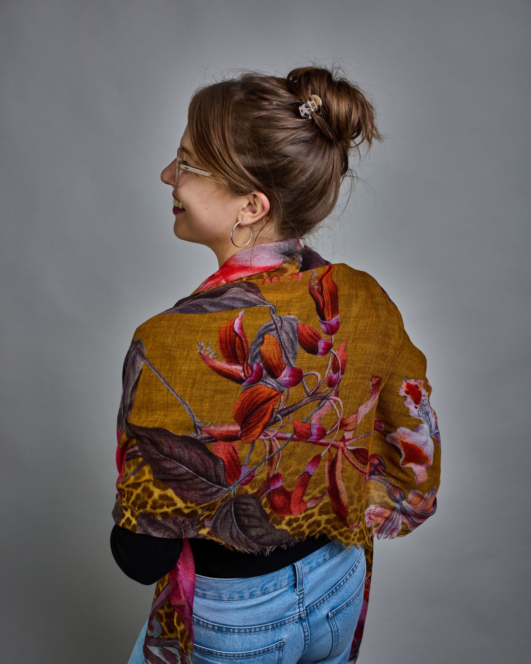 Ein Schal aus 75 % Wolle und 25 % Modal mit den Maßen 70 × 180 cm. Je nach Bild ist er locker um den Nacken gelegt oder einmal gewickelt, um eine lockere Schlaufe um den Hals zu haben, oder zweimal gewickelt, um das Maximum an Wärme zu bekommen. An den Kanten ist er leicht gefranst. Die Hintergrundfarbe des Schals ist Okker und hier und da ist ein Leomuster in Brauntönen zu sehen. Auf dem ganzen Schal sind Blumen in Rot-, Orange-, Weiß- und Pinktönen sowie Blätter beziehungsweise Blumenstängel in verschied…