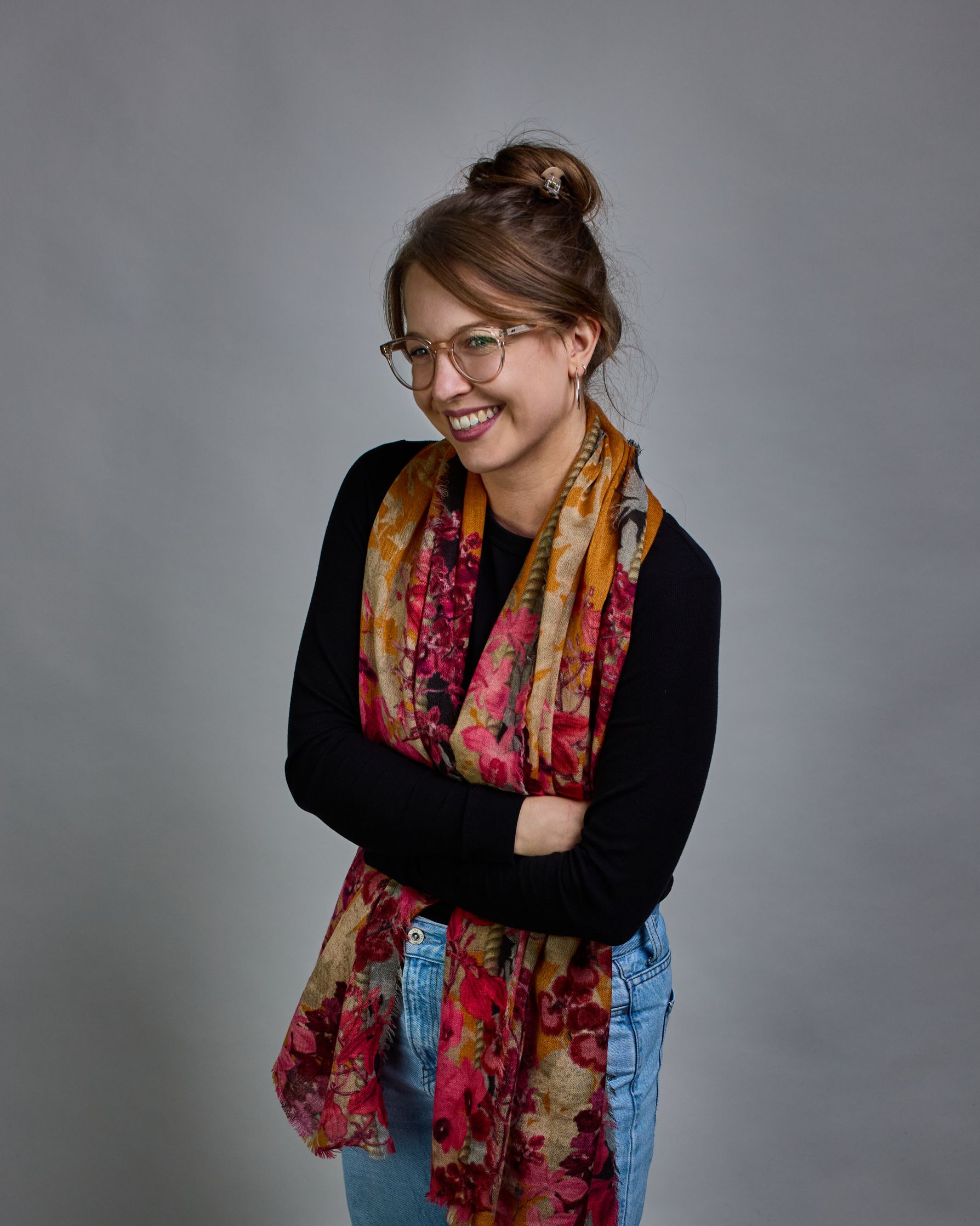 Ein Schal aus 75 % Wolle und 25 % Modal mit den Maßen 70 × 180 cm. Je nach Bild ist er locker um den Nacken gelegt oder einmal gewickelt, um eine lockere Schlaufe um den Hals zu haben, oder zweimal gewickelt, um das Maximum an Wärme zu bekommen. An den Kanten ist er leicht gefranst. Die Hintergrundfarbe des Schals ist Okker. Von den schmalen Kanten des Schals kommen die Blumen in Richtung Mitte. Die Blüten sind in Rosa-, Lila- und Weißtönen gestaltet. #1